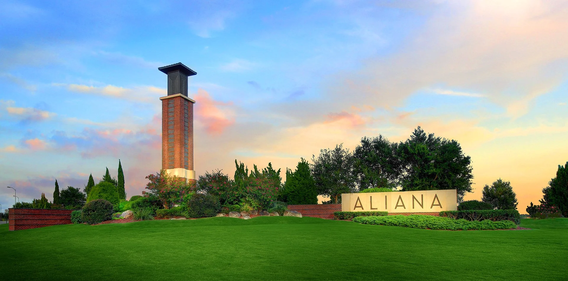 Community entry sign that reads 'ALHANA' near a lush green lawn, trees, and shrubs, with a brick tower and a colorful sky with clouds at sunset in the background.