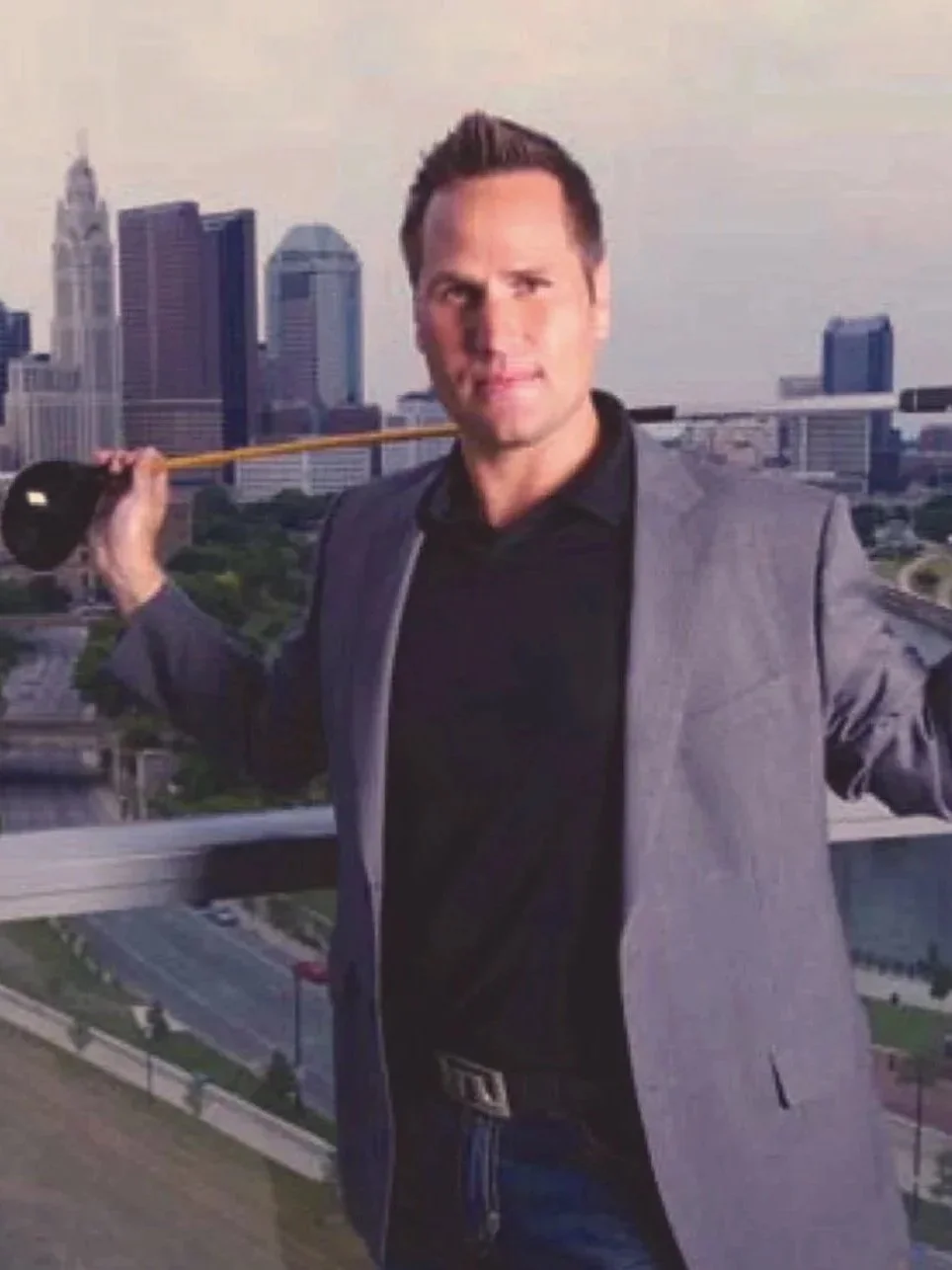 A man holding a golf club over his shoulders on a rooftop with a city skyline in the background.