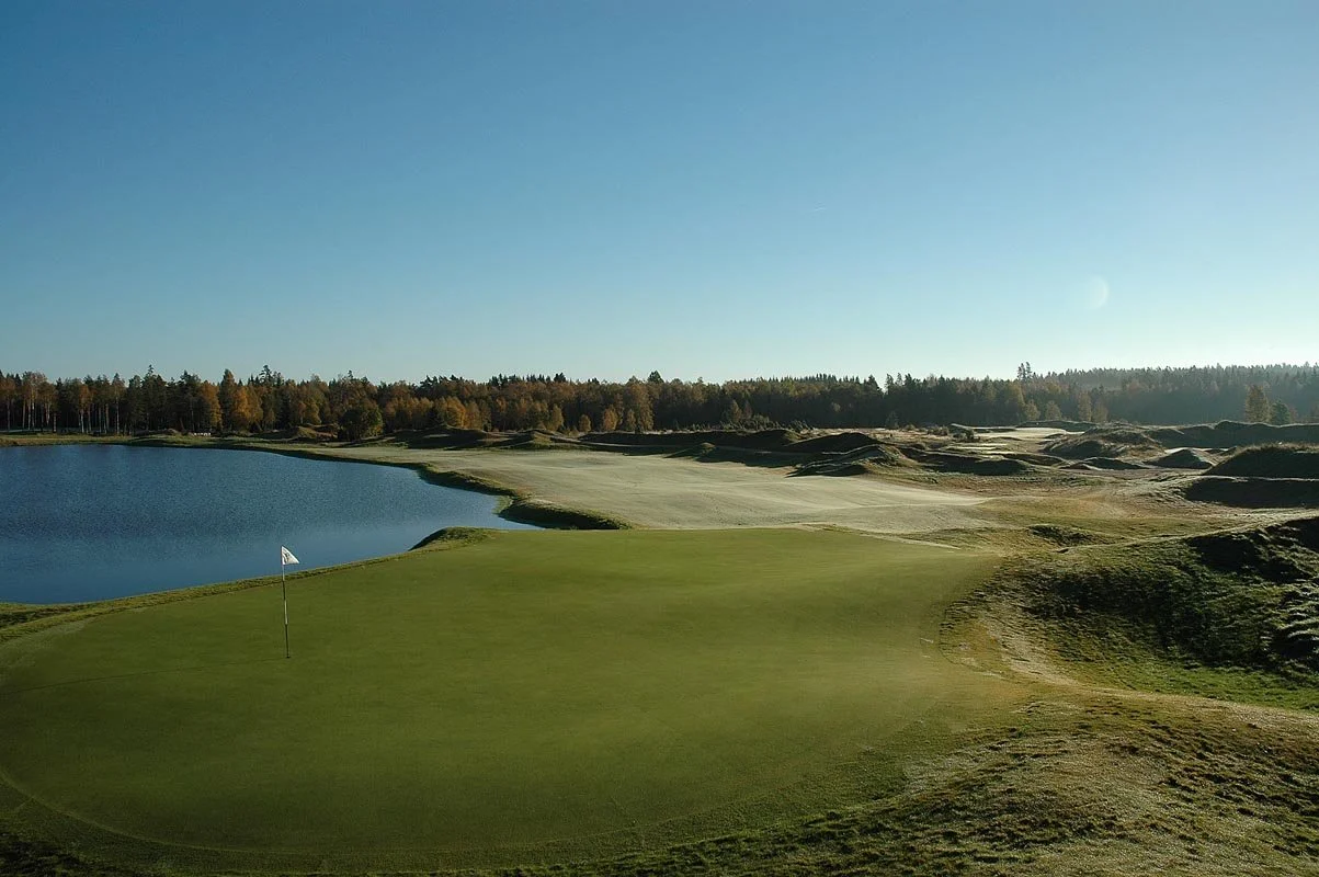 A scenic golf course with a green, a flagstick, a water hazard, and rolling terrain, surrounded by trees under a clear sky.
