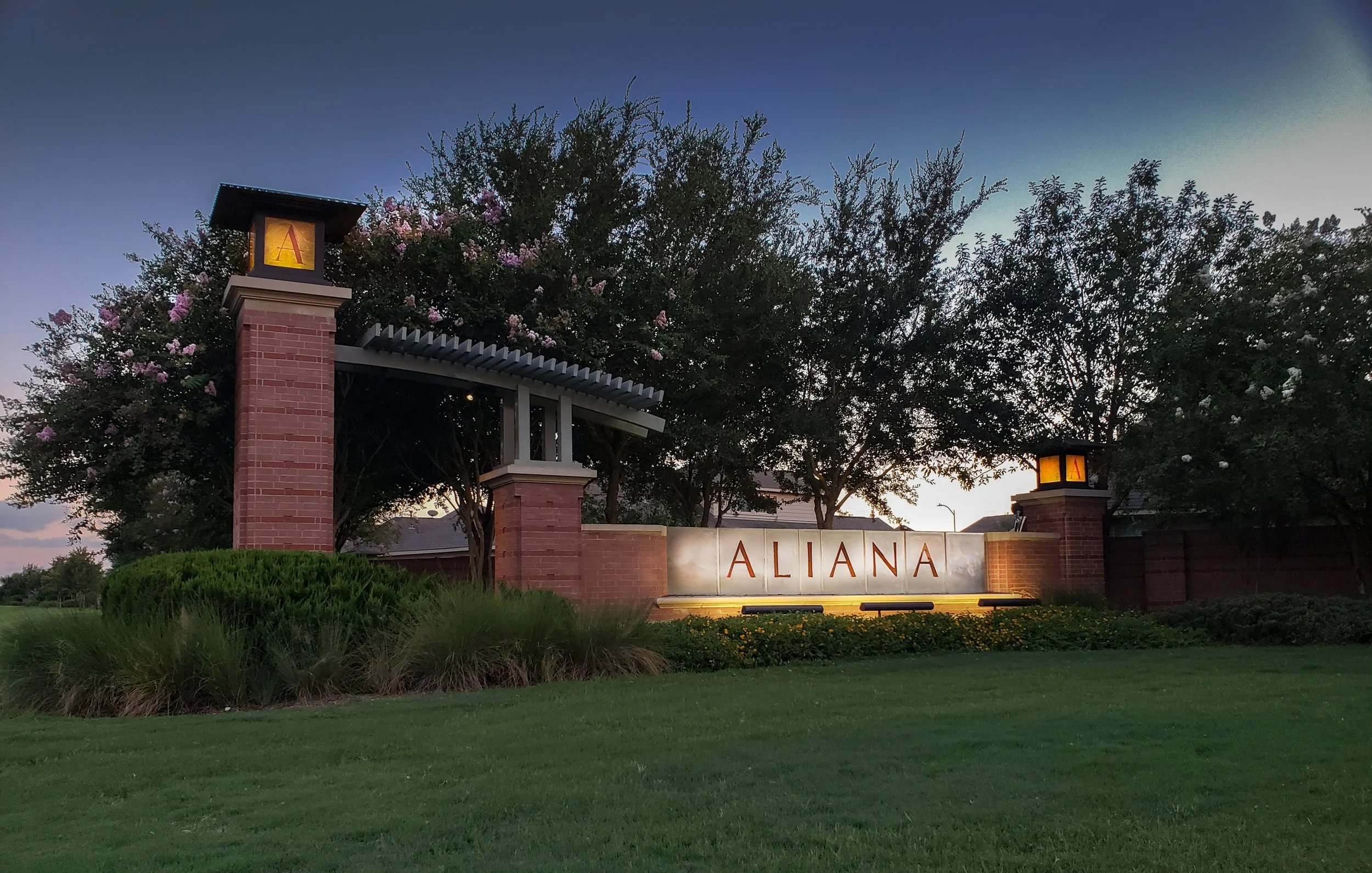 Entrance sign to Aliana neighborhood with illuminated pillars, trees, and landscaping at dusk.