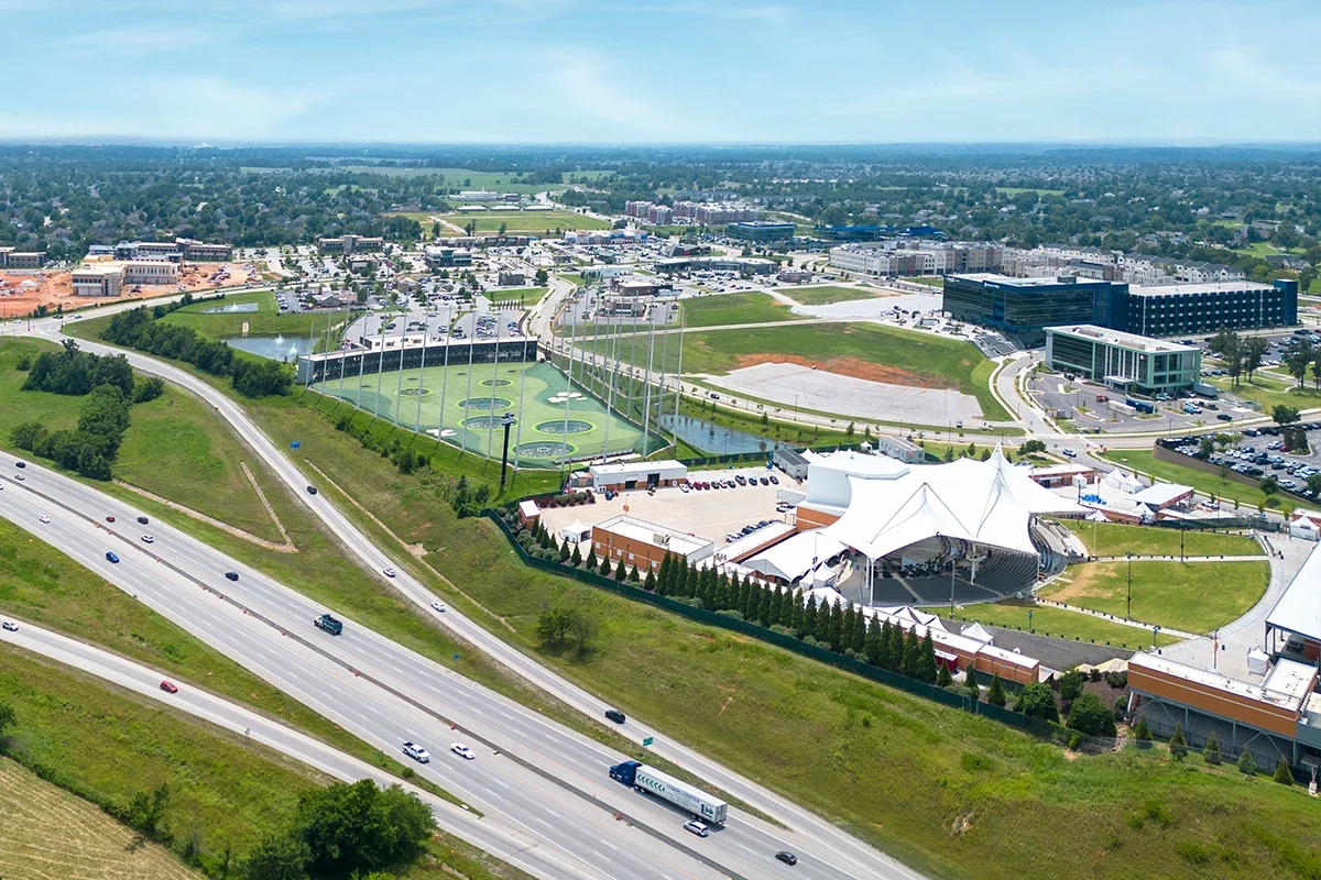 Aerial view of a mixed-use development with multiple buildings, parking lots, a golf practice range, a white tented event space, amphitheater and nearby highways, with green fields and residential areas in the distance.