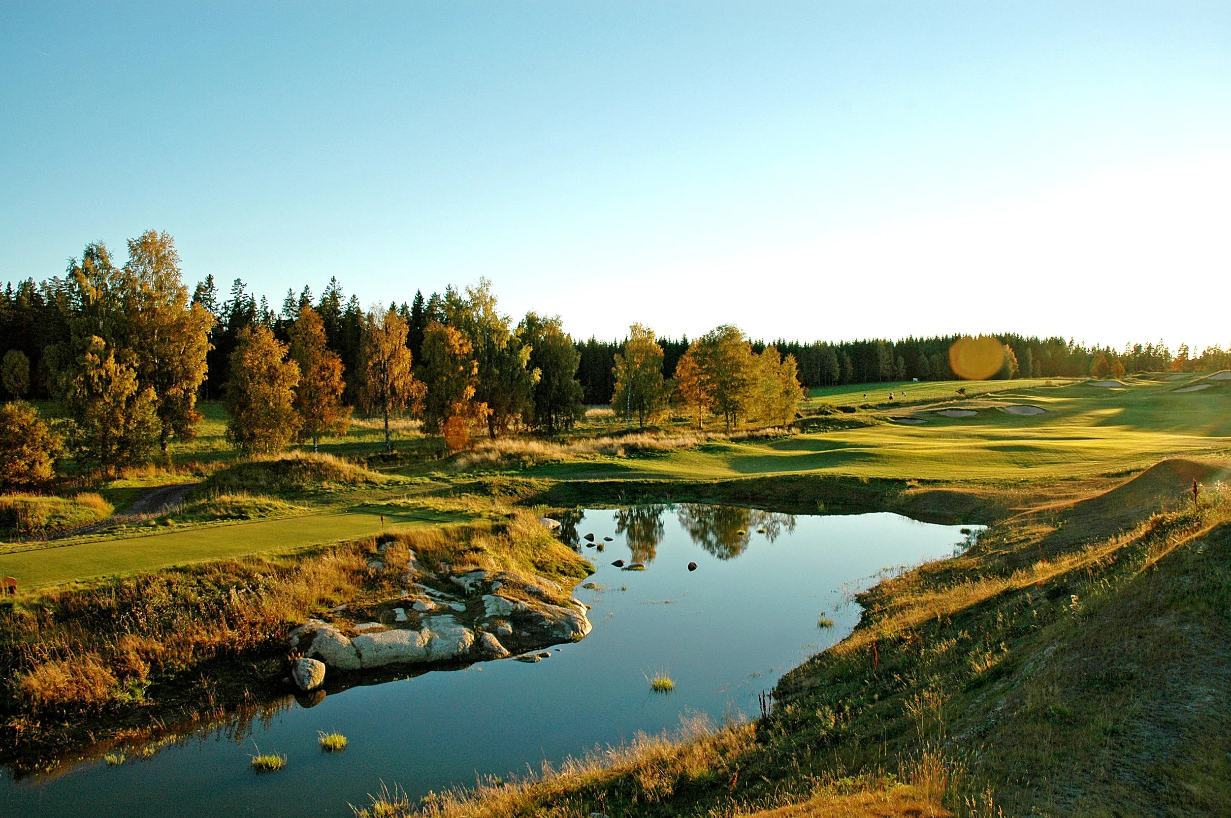 A scenic golf course with green fairways, a small pond reflecting the sky, and autumn-colored trees under a clear sky.