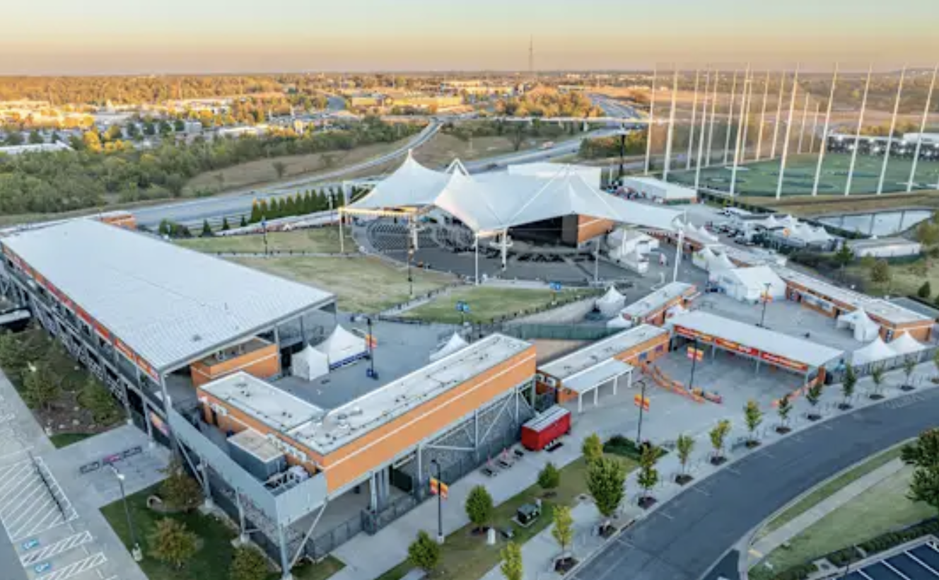 Aerial view of a modern mixed-use entertainment complex with a large tented roof structure, multiple smaller buildings, parking lots, and green spaces at sunset.