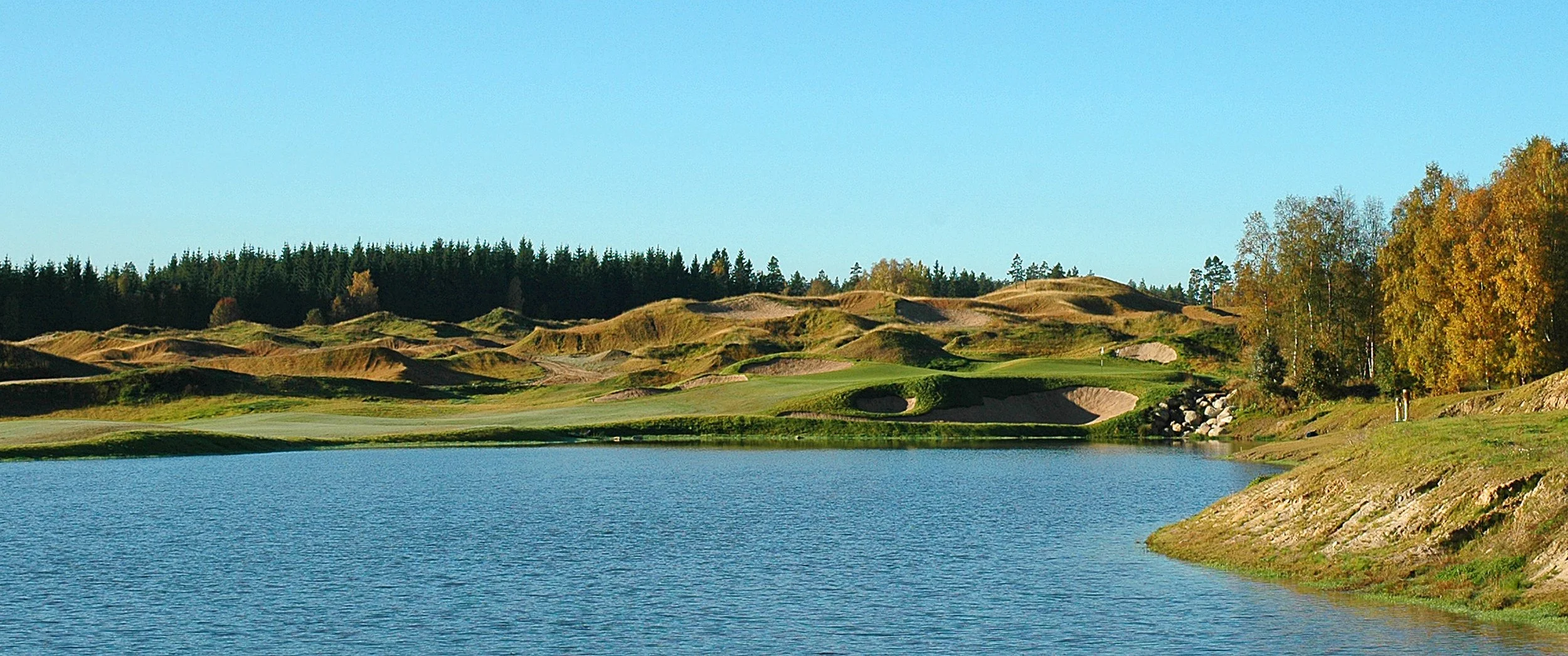 A landscape of a golf course with a water hazard, sand traps, rolling hills, and trees during daytime.