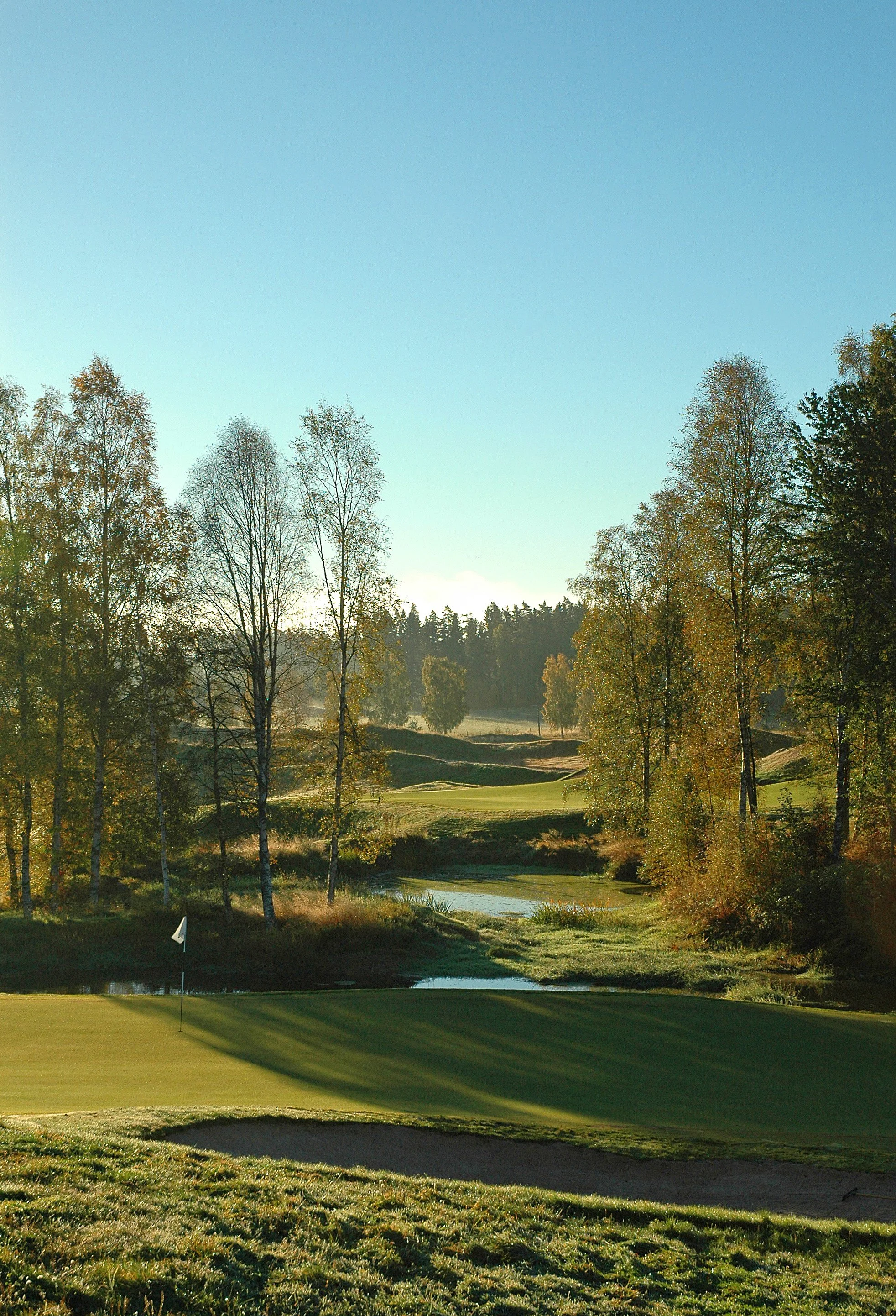 A scenic golf course with a green in the foreground, trees with fall foliage, and a small water feature in the middle distance, under a clear blue sky.