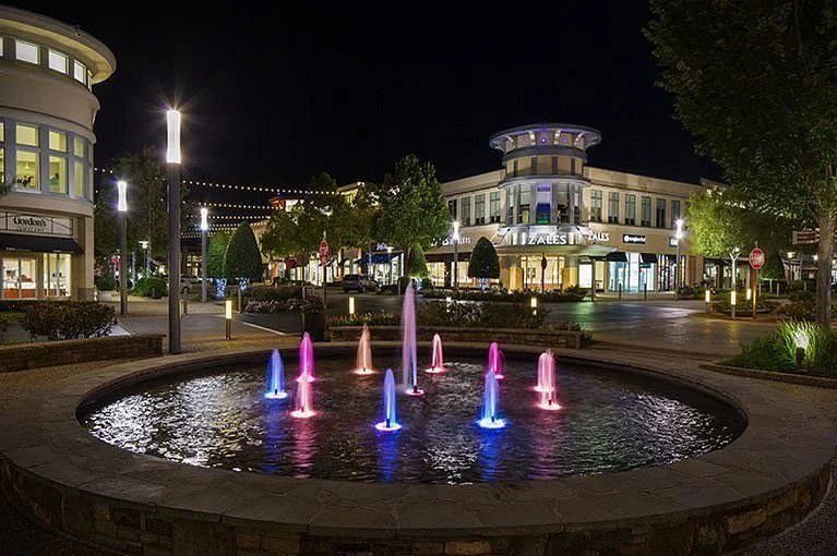 Night scene of a shopping area with a lit fountain featuring colorful lights in the foreground, surrounded by trees and modern buildings.