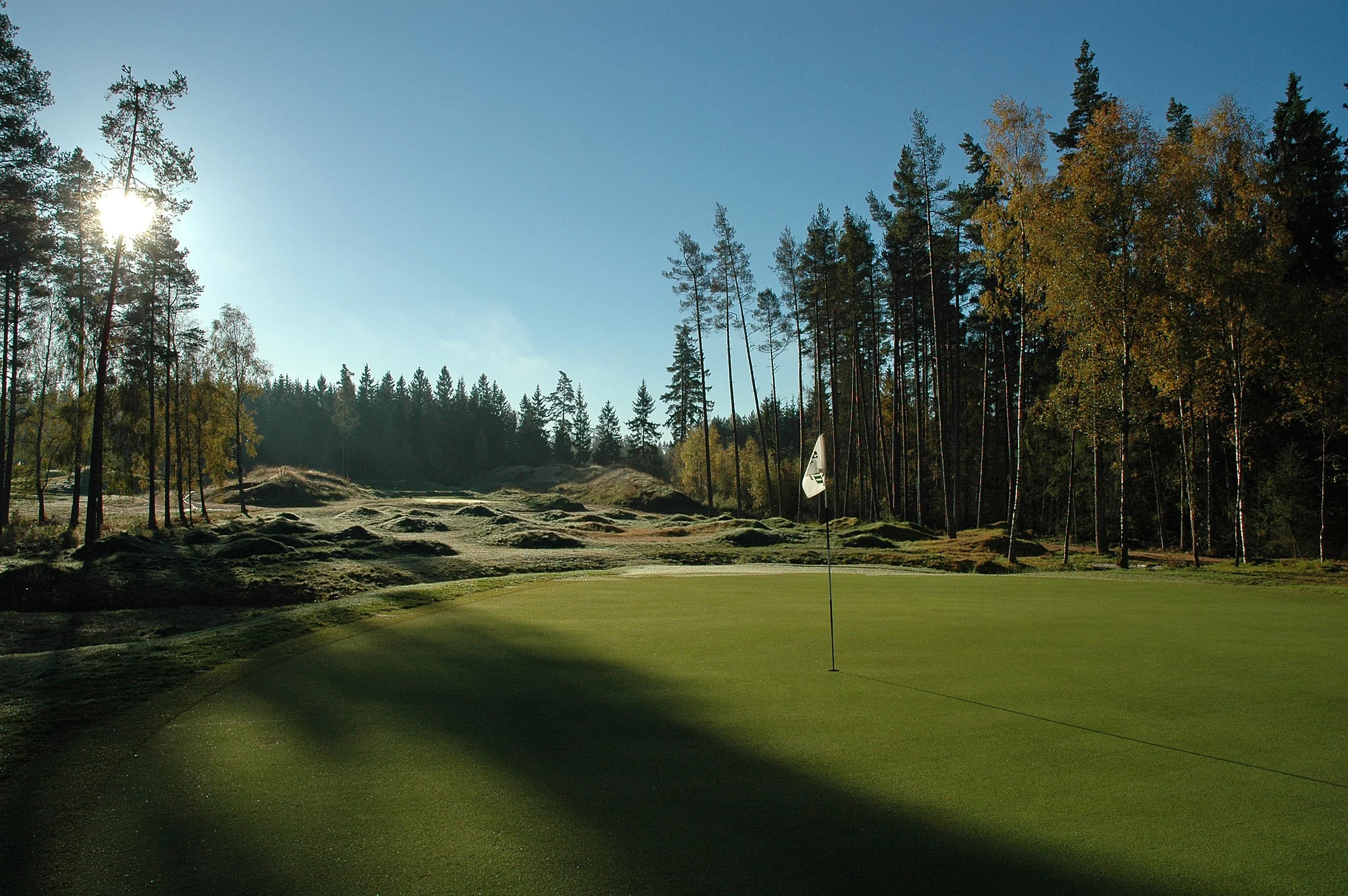 A golf course with a putting green, flagstick, surrounding dirt and grass, and tall trees under a clear blue sky with sunlight shining through.