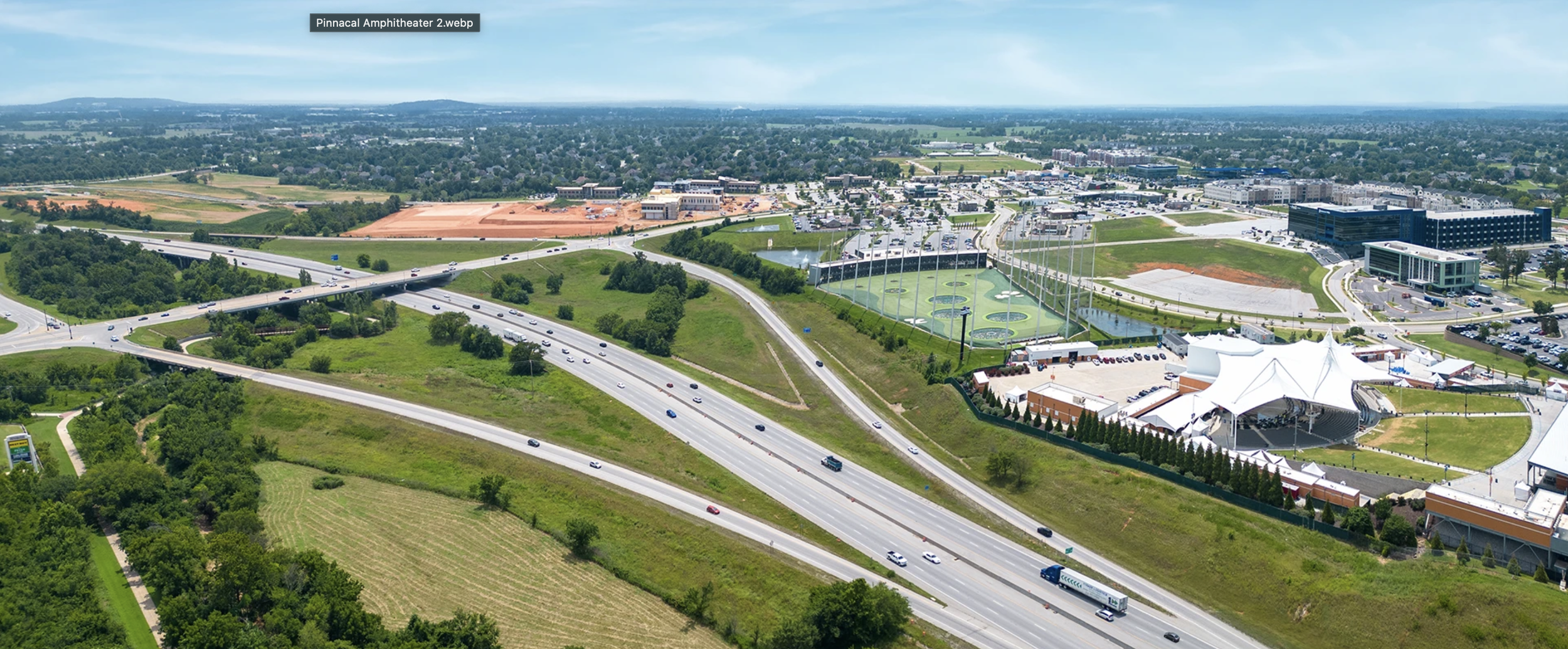 Aerial view of Pinnacle Amphitheater and surrounding area with roads, parking lots, buildings, green spaces, golf driving range, and a large stage area with a white canopy tent.