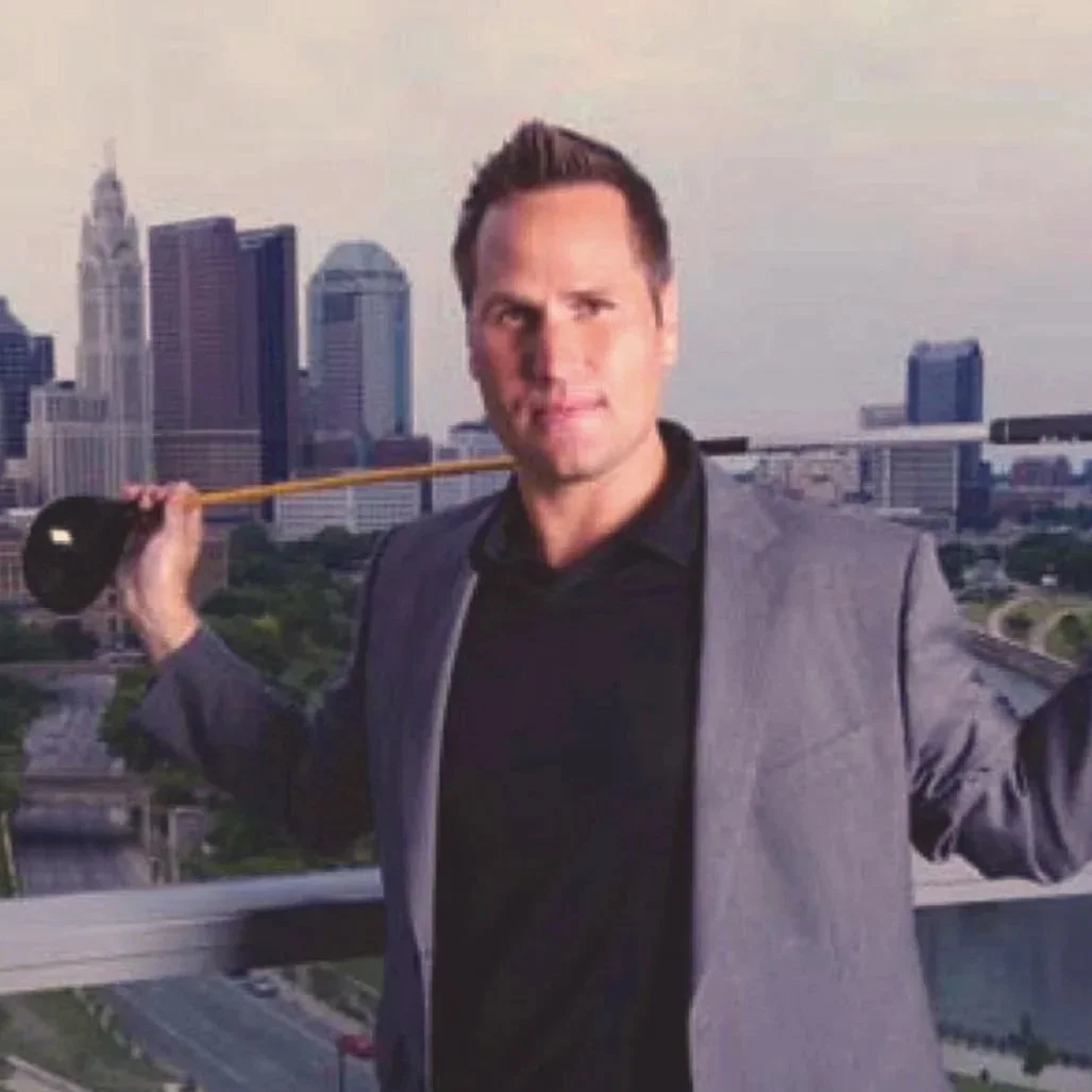 A man in a gray suit holding a golf club over his shoulder on a rooftop with a city skyline in the background.