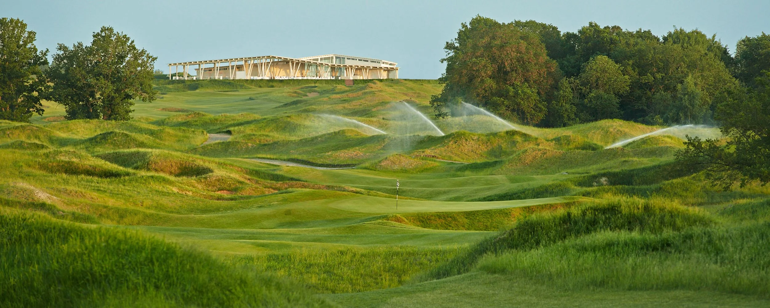 A golf course with a green, flags, sand bunkers, and water sprinklers, surrounded by trees and a contemporary building in the background.