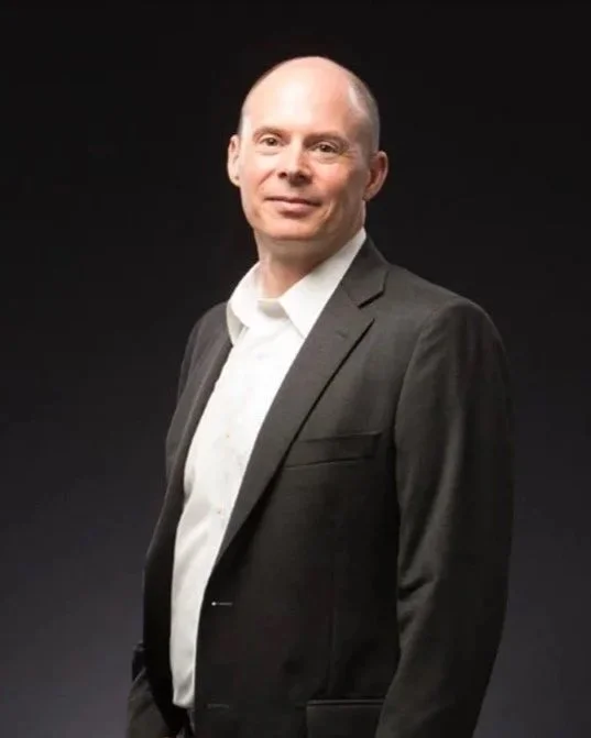 A bald man wearing a white shirt and a dark suit jacket, standing against a dark background, looking at the camera with a slight smile.