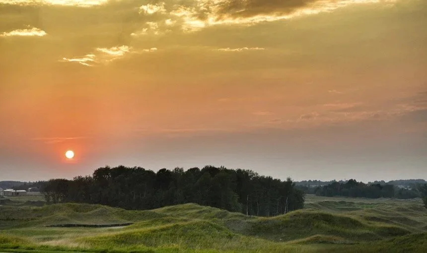 Sunset over rolling green hills with a forested area and a sky with orange and yellow hues.
