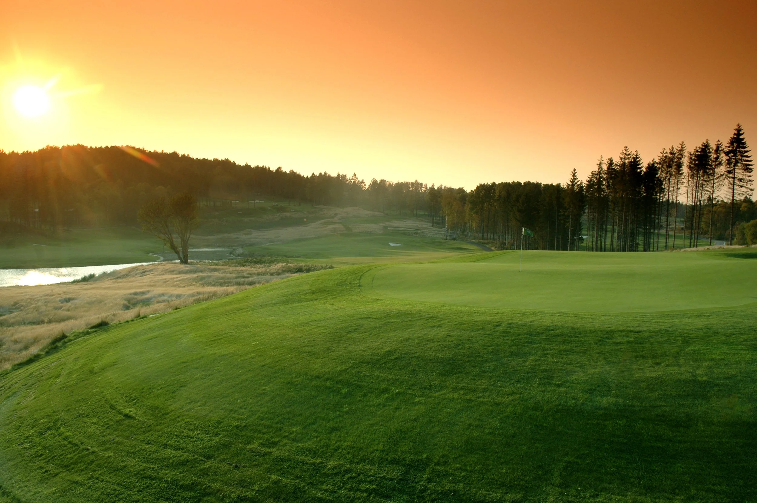 A golf course at sunset with green grass, a few trees, and a flag on the green. The sky is orange and there are tall trees in the background.