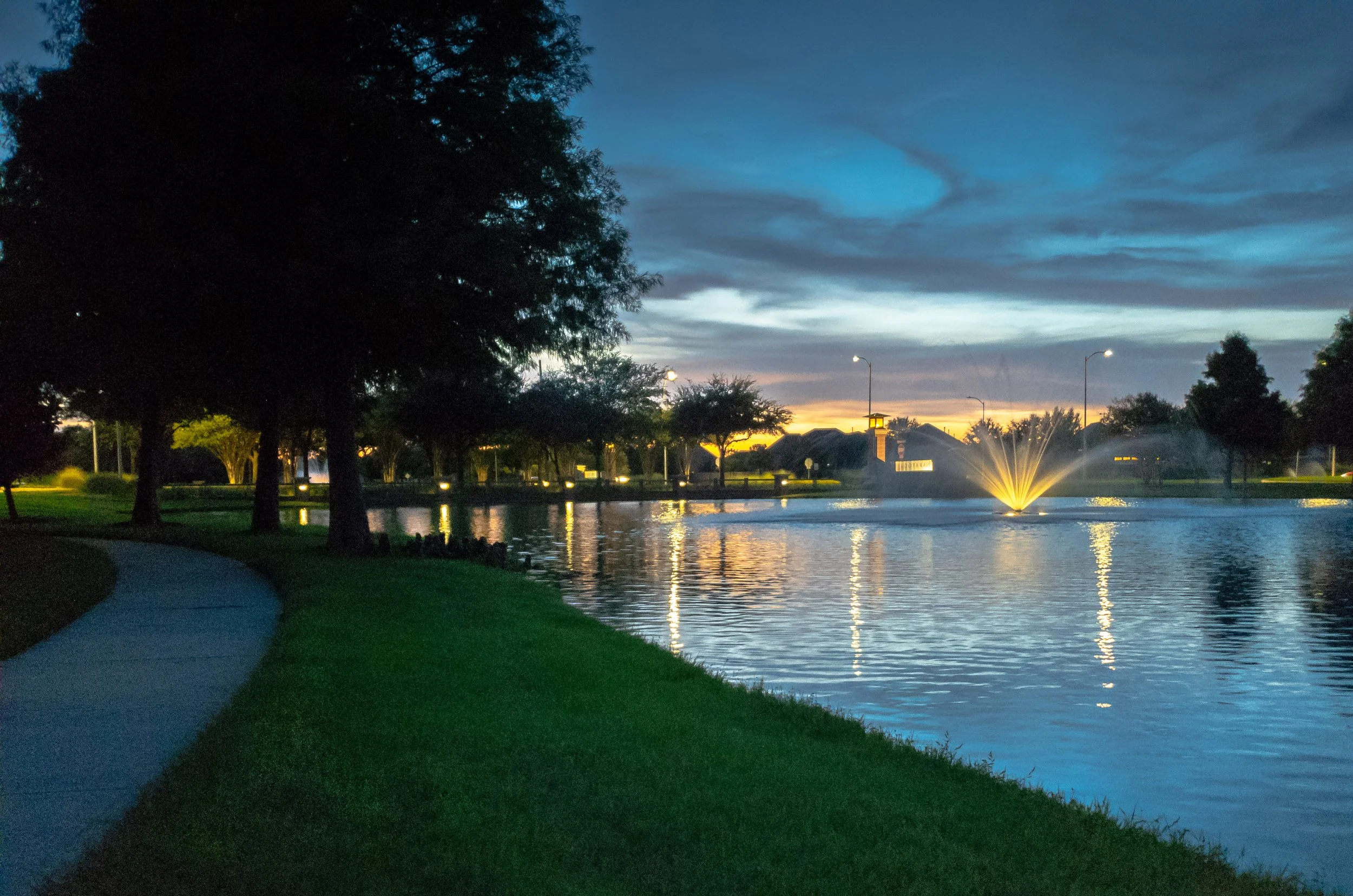 A park during sunset with a pond, trees, a fountain, and streetlights illuminating the area.