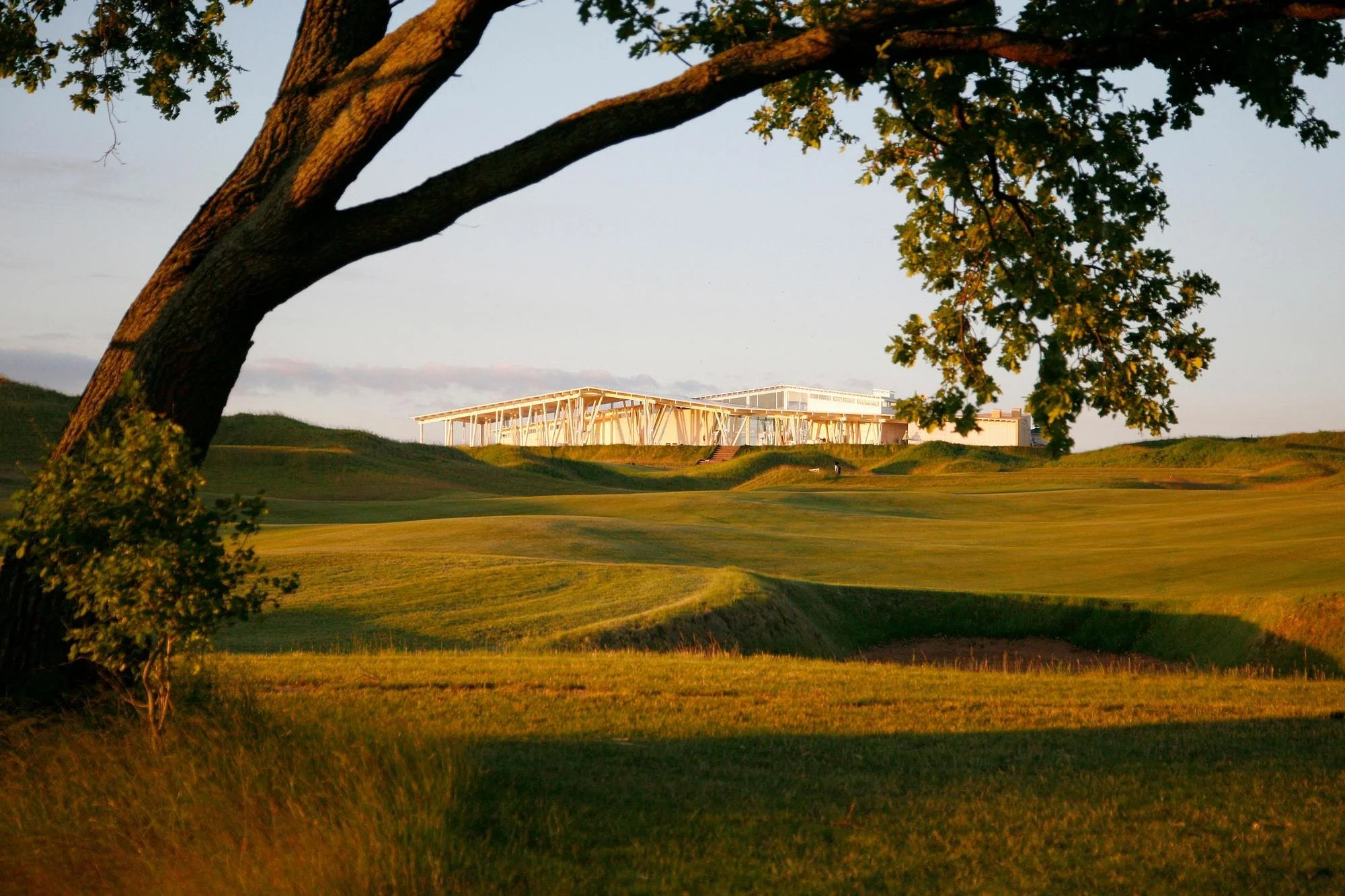 A golf course at sunset with lush green grass, a large tree in the foreground, and a modern building with white structural elements and glass windows in the background.