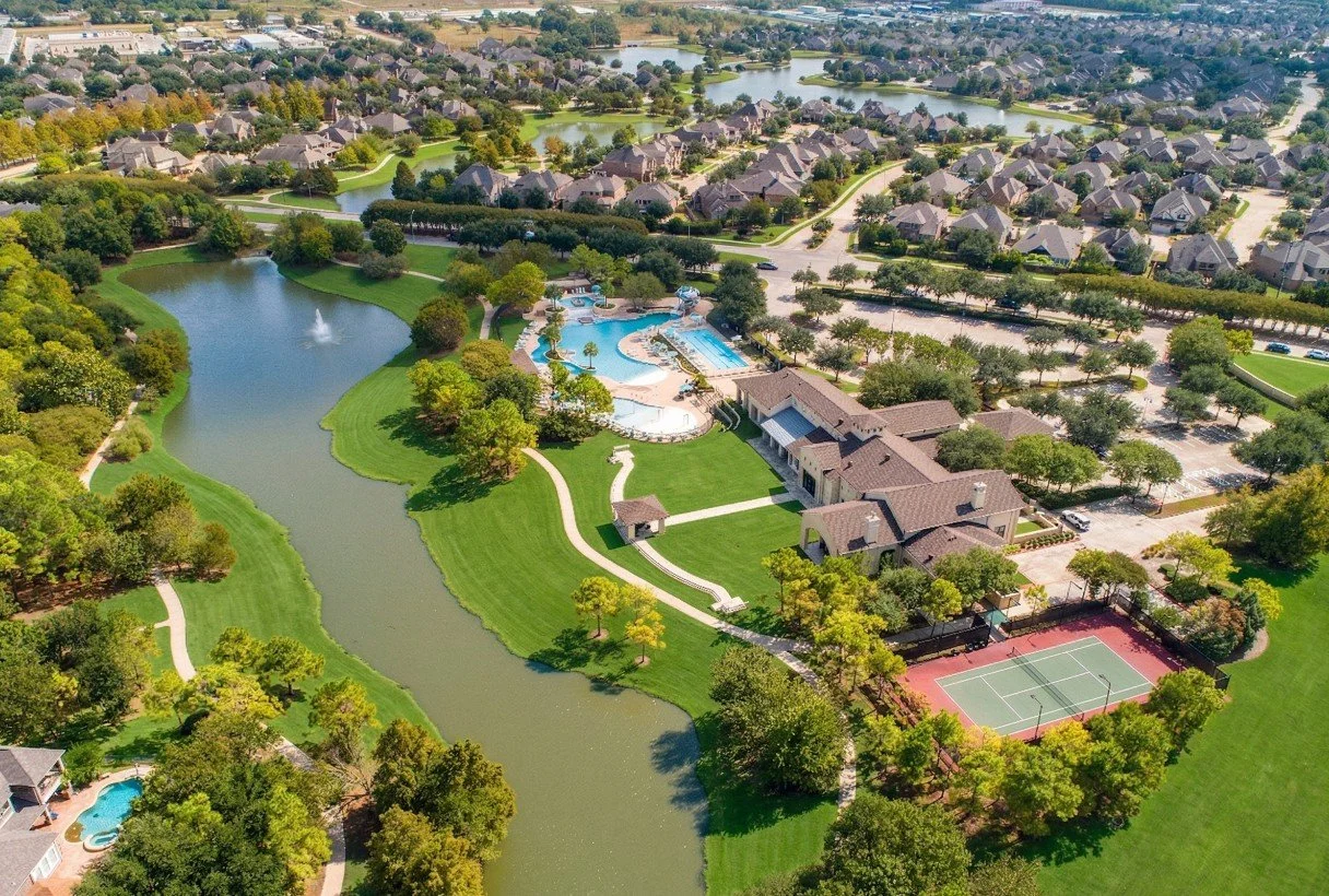 Aerial view of a residential community with a park, pond, swimming pool, tennis court, and well-maintained houses surrounded by trees.
