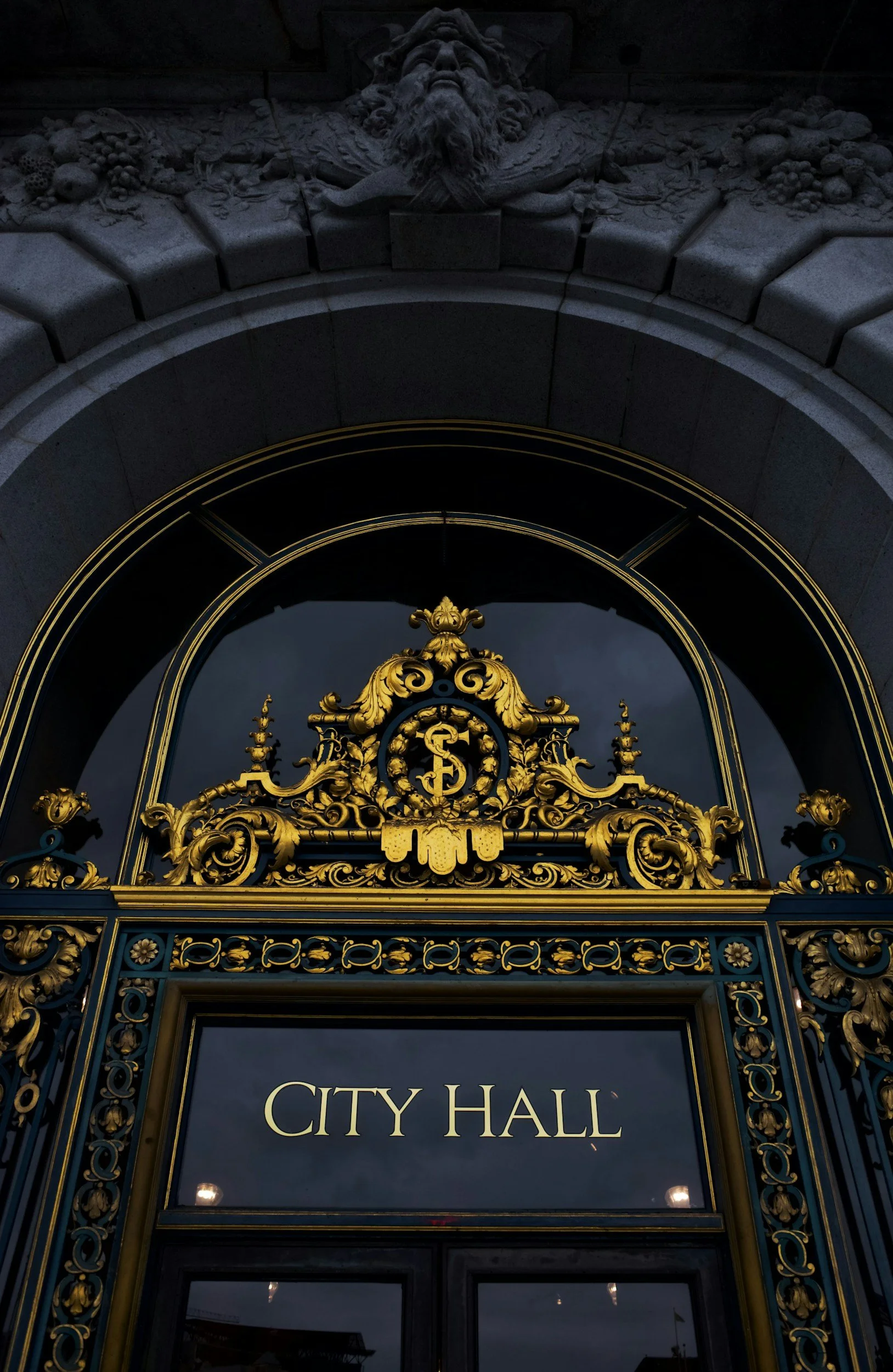 The entrance to a city hall building with ornate gold and teal ironwork framing a glass door that has 'City Hall' written on it, unlit.