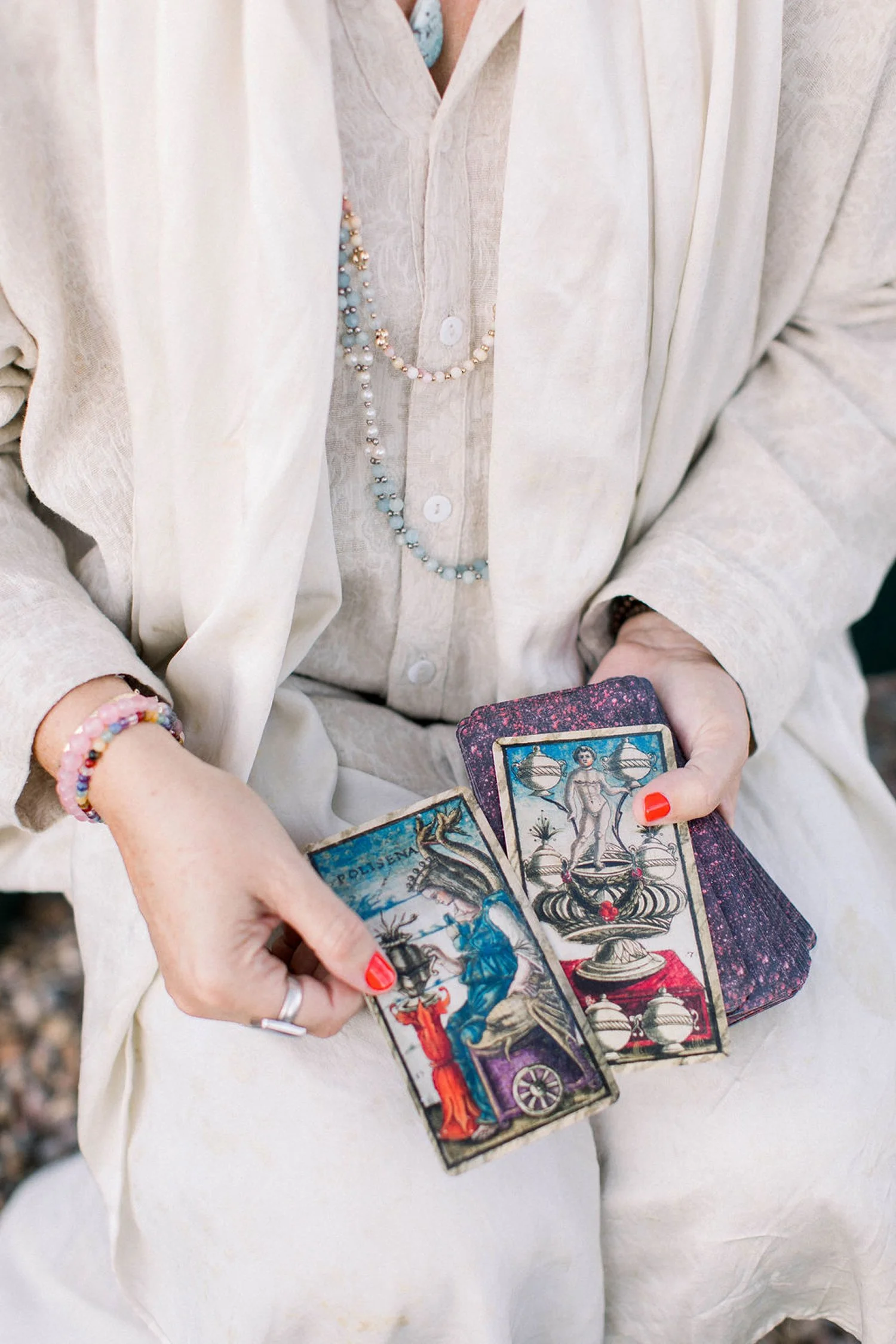A person dressed in light-colored clothing holding tarot cards and wearing jewelry, including necklaces and bracelets.