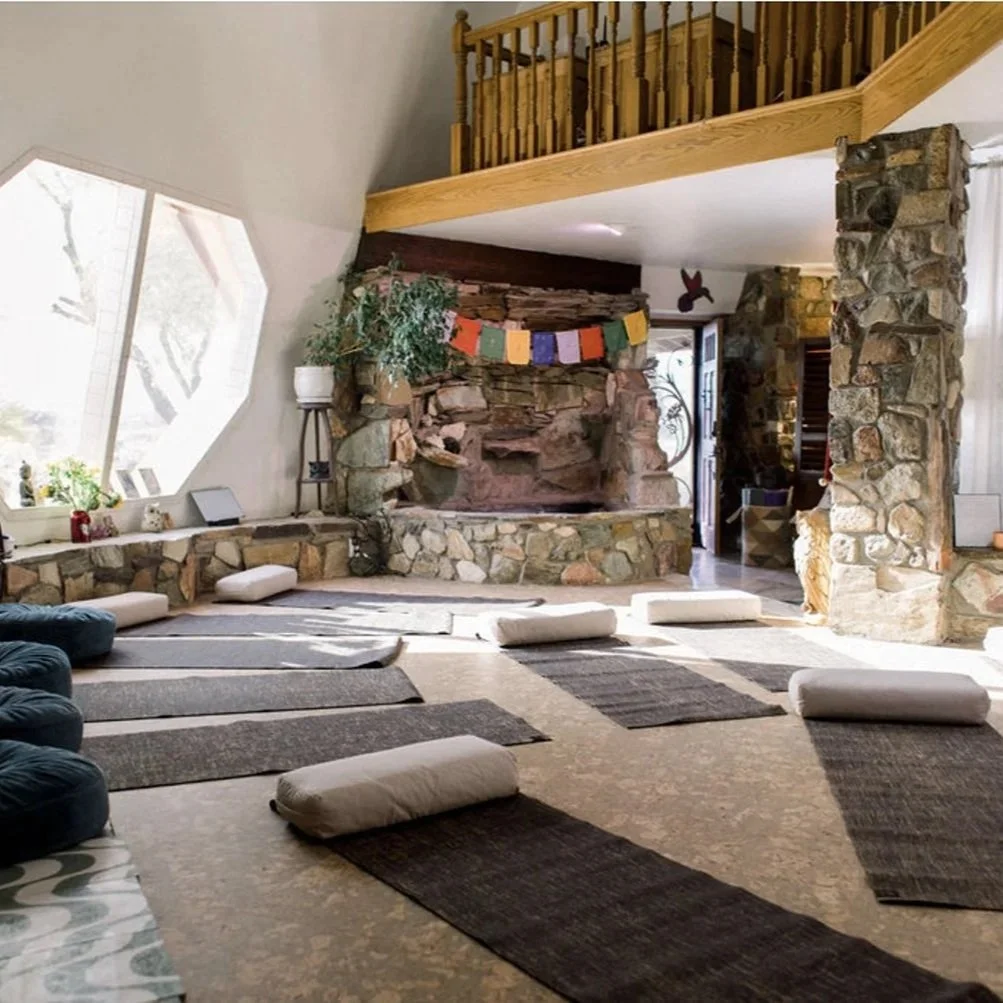 Sunlit interior room with mats and cylindrical cushions on the floor, stone fireplace decorated with colorful prayer flags, large window with plants, and wooden balcony with railing overhead.