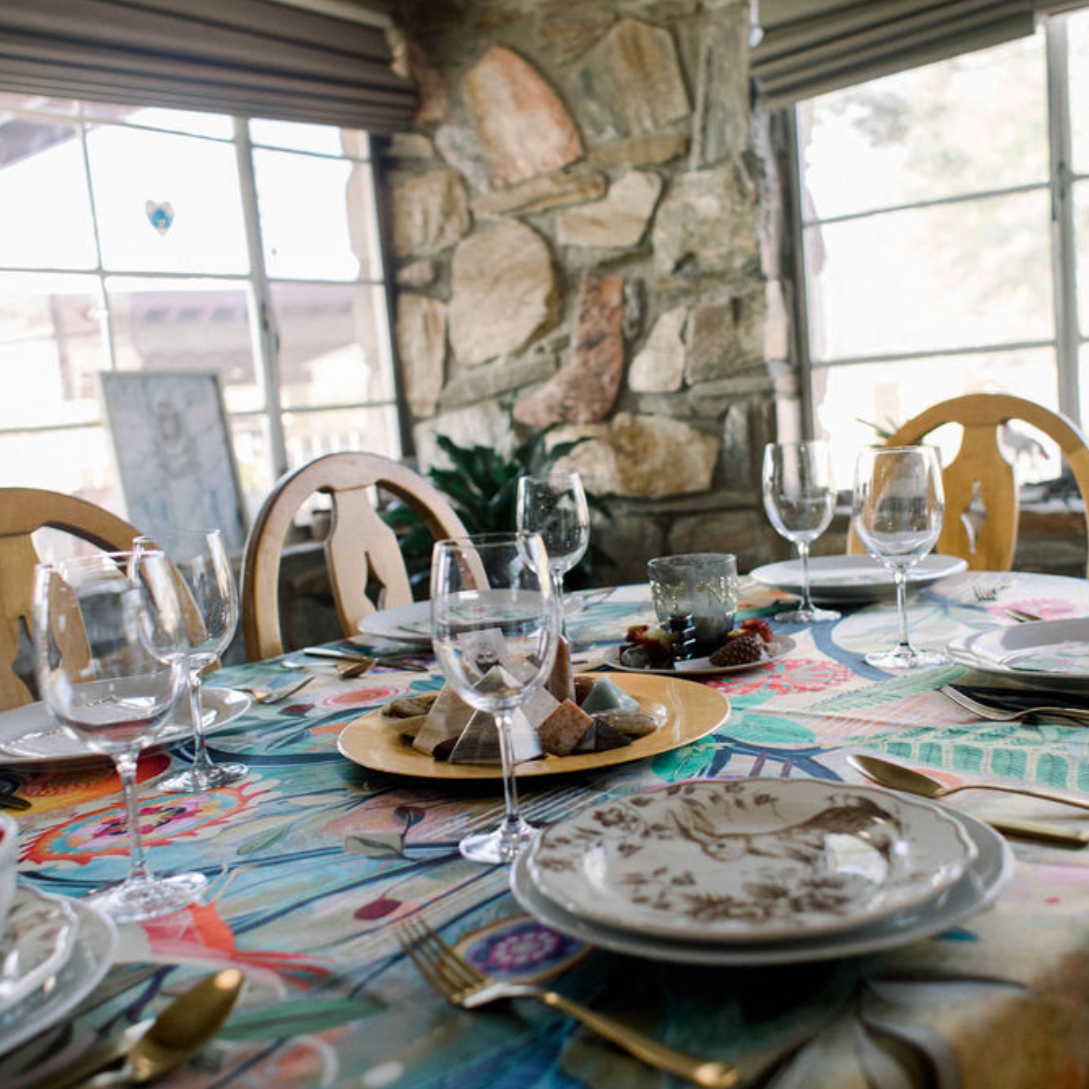 A dining table set with plates, wine glasses, and gold-colored utensils inside a room with large windows and a stone wall.