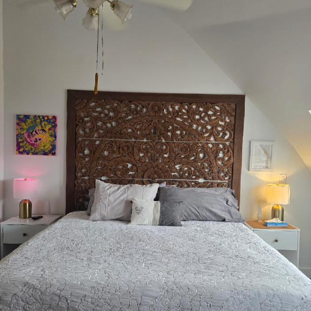 A bedroom with a bed that has a large, intricately carved wooden headboard, white floral quilt, and multiple pillows. There are two white nightstands with lamps on each side, one with a pink lamp and the other with a yellow lamp, and colorful artwork hanging on the walls.