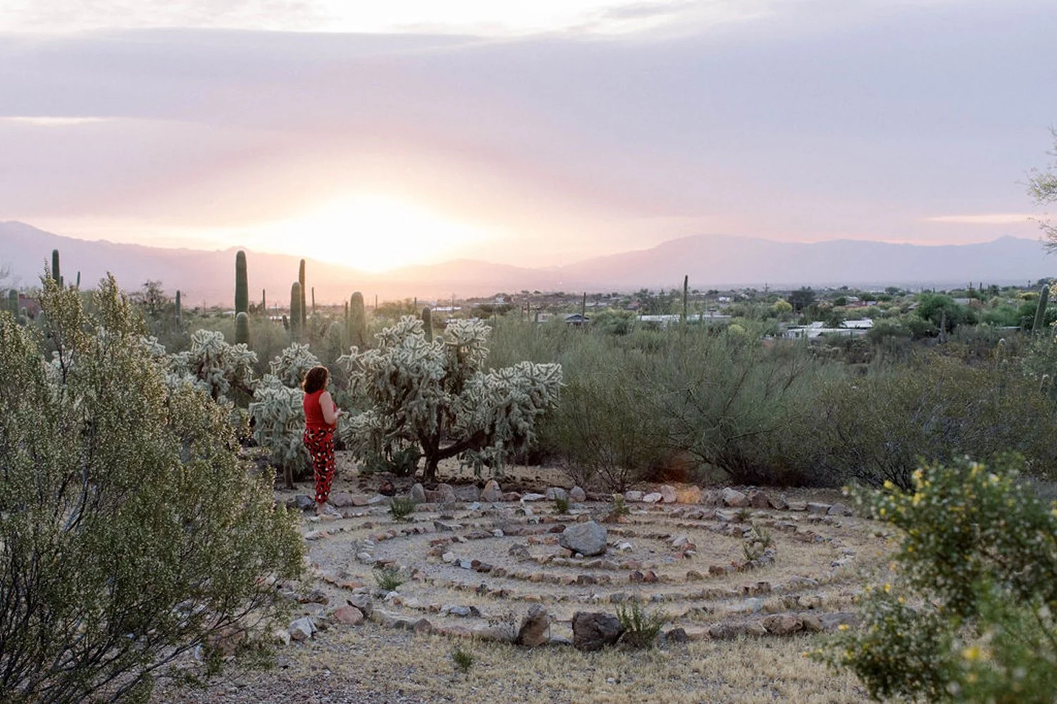 A woman in red and black clothing walks a labyrinth stone circle in a desert garden at sunset, surrounded by cacti and desert shrubs, with mountains in the distance.