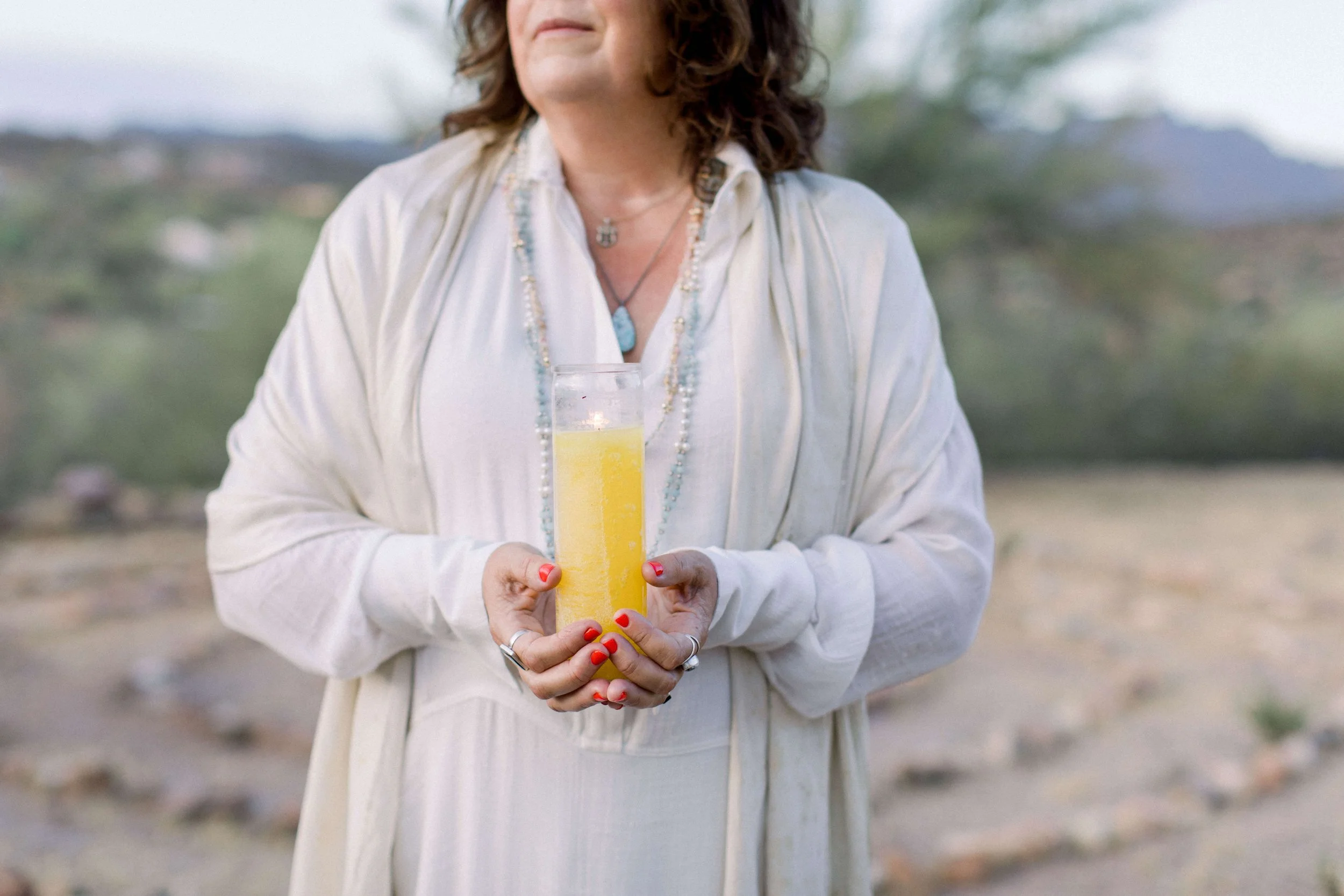 A woman with curled hair wearing white robes and layered necklaces holding a tall glass of yellow drink outdoors, with a blurred natural background.