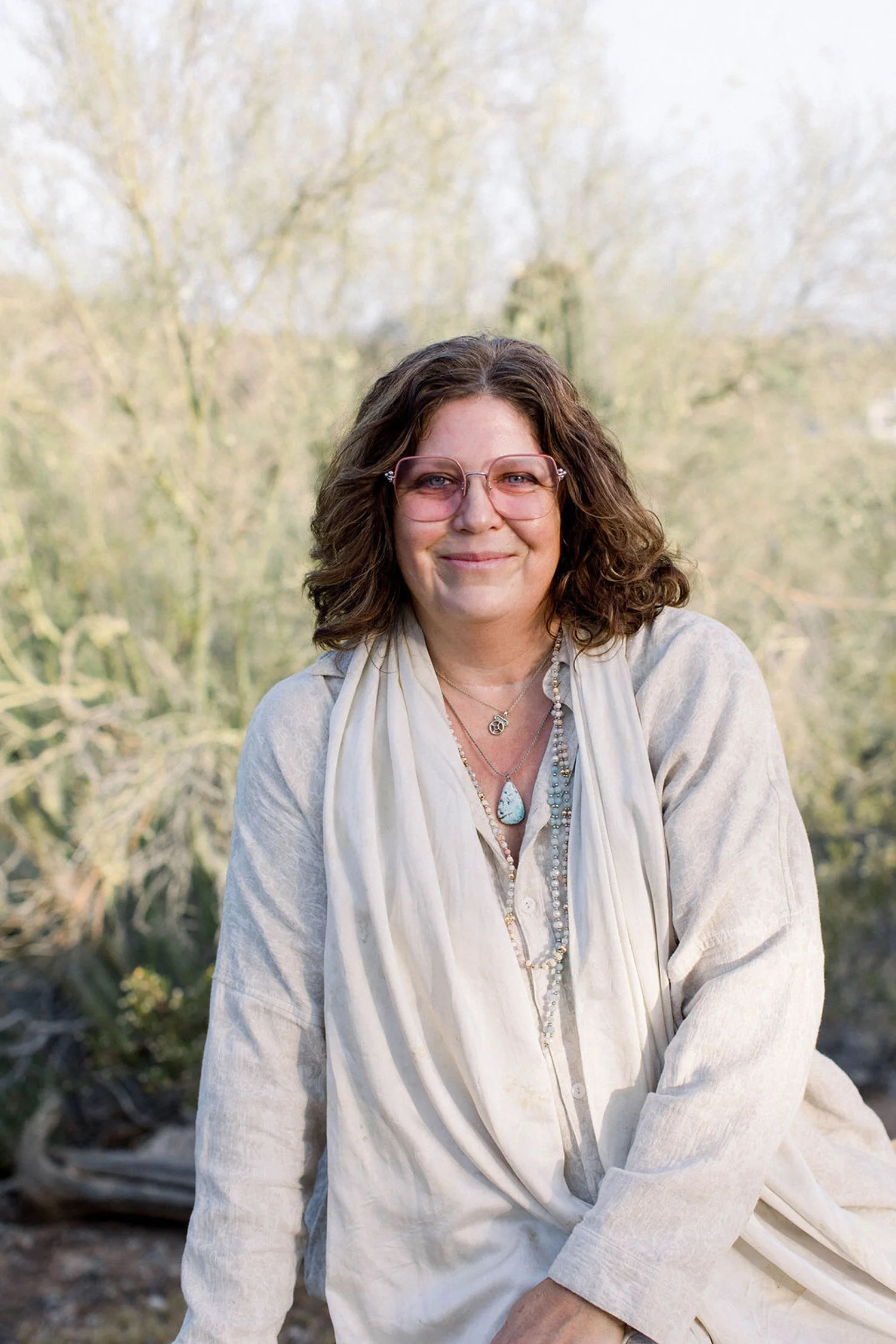 A woman with curly brown hair and wearing pink glasses, a light-colored shirt, and layered necklaces, sitting outdoors with a blurred natural background of trees in early spring.
