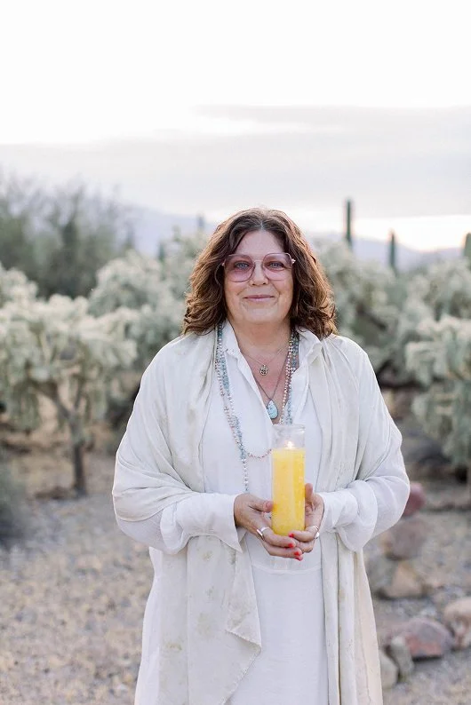 A woman with curly brown hair and glasses standing outdoors in a desert landscape with cacti in the background. She is wearing a white dress and a light-colored jacket, holding a glass with a yellow beverage. She has necklaces around her neck and a gentle smile.