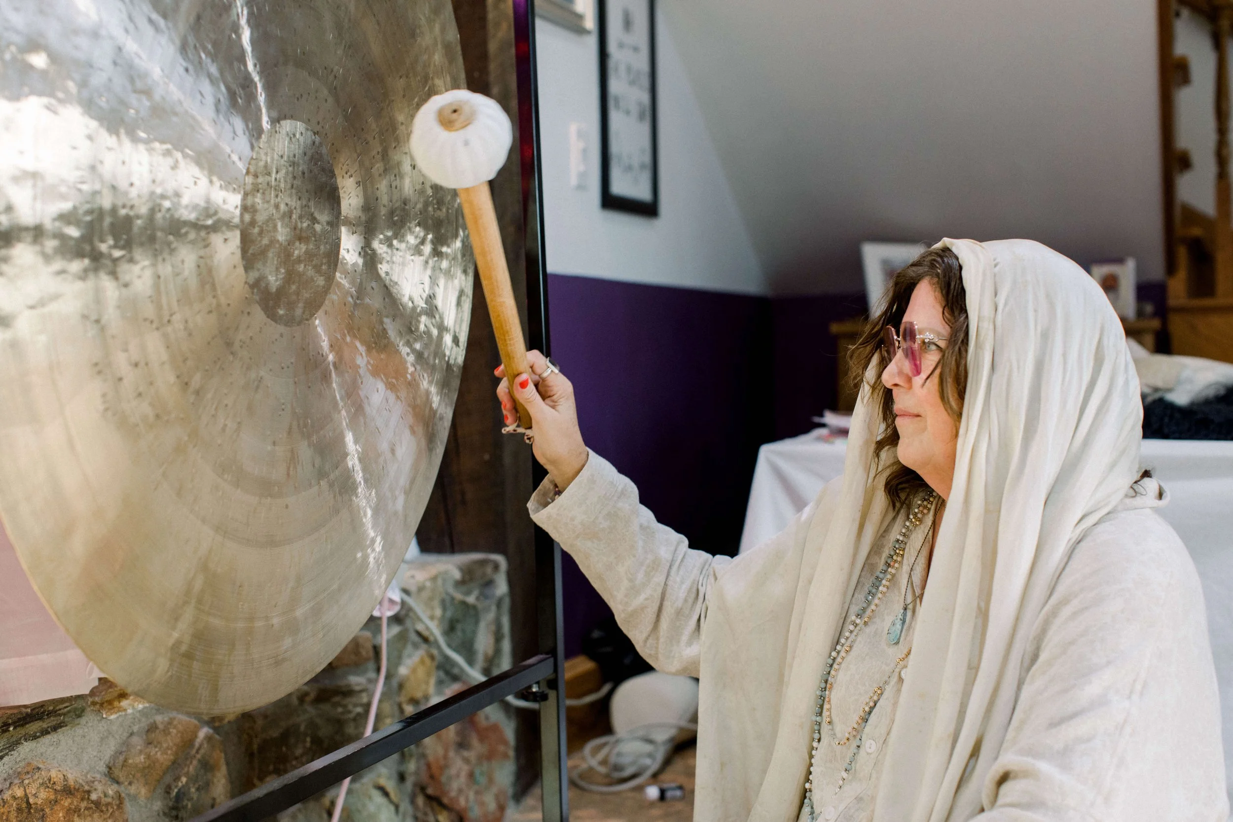 A woman dressed in white with a head covering and glasses is striking a gong with a mallet. The gong is large and gold-colored, with a person’s reflection visible on its surface. The setting appears to be indoors, possibly in a home or studio.
