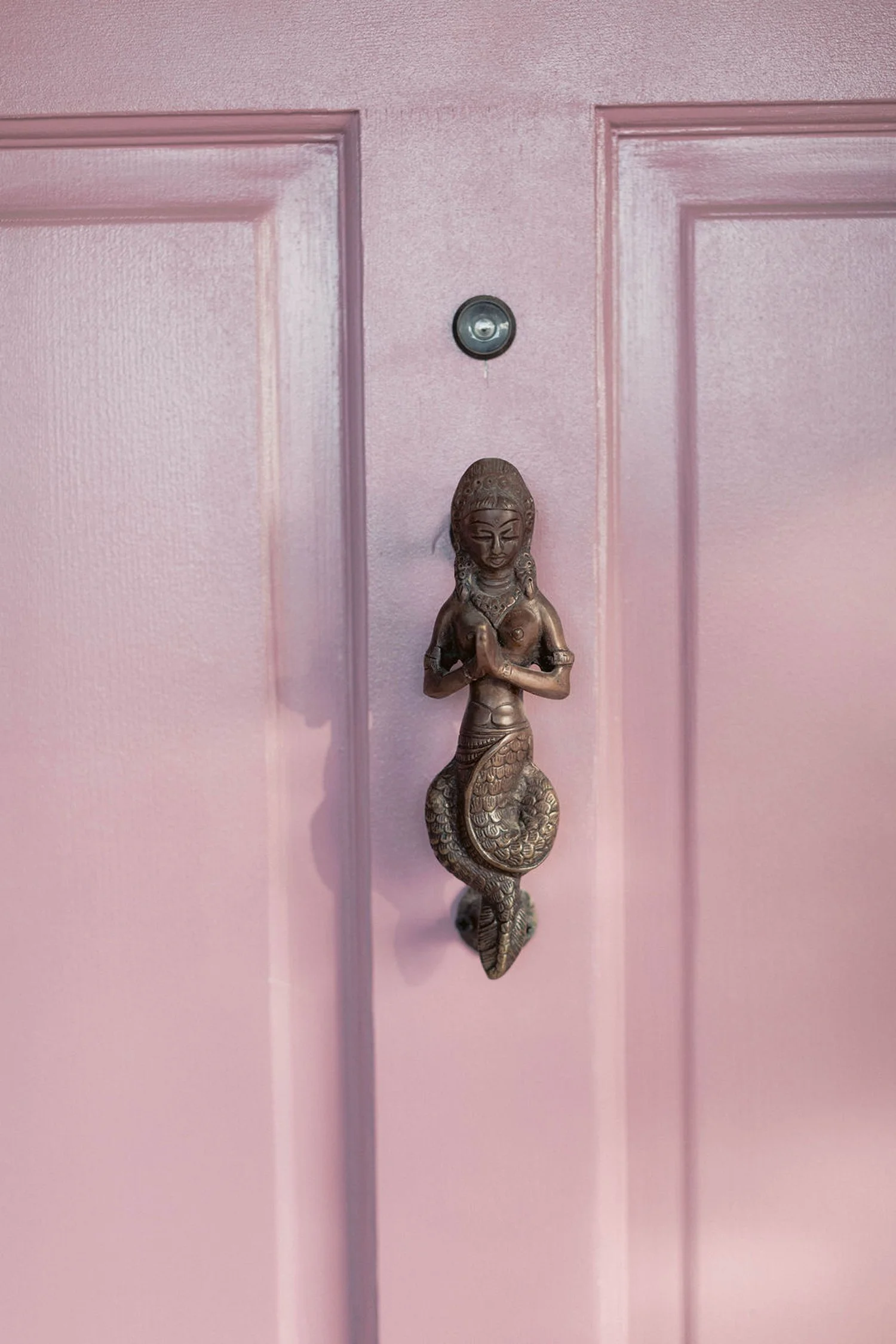 A decorative door knocker shaped like a mermaid, mounted on a pink-colored door.