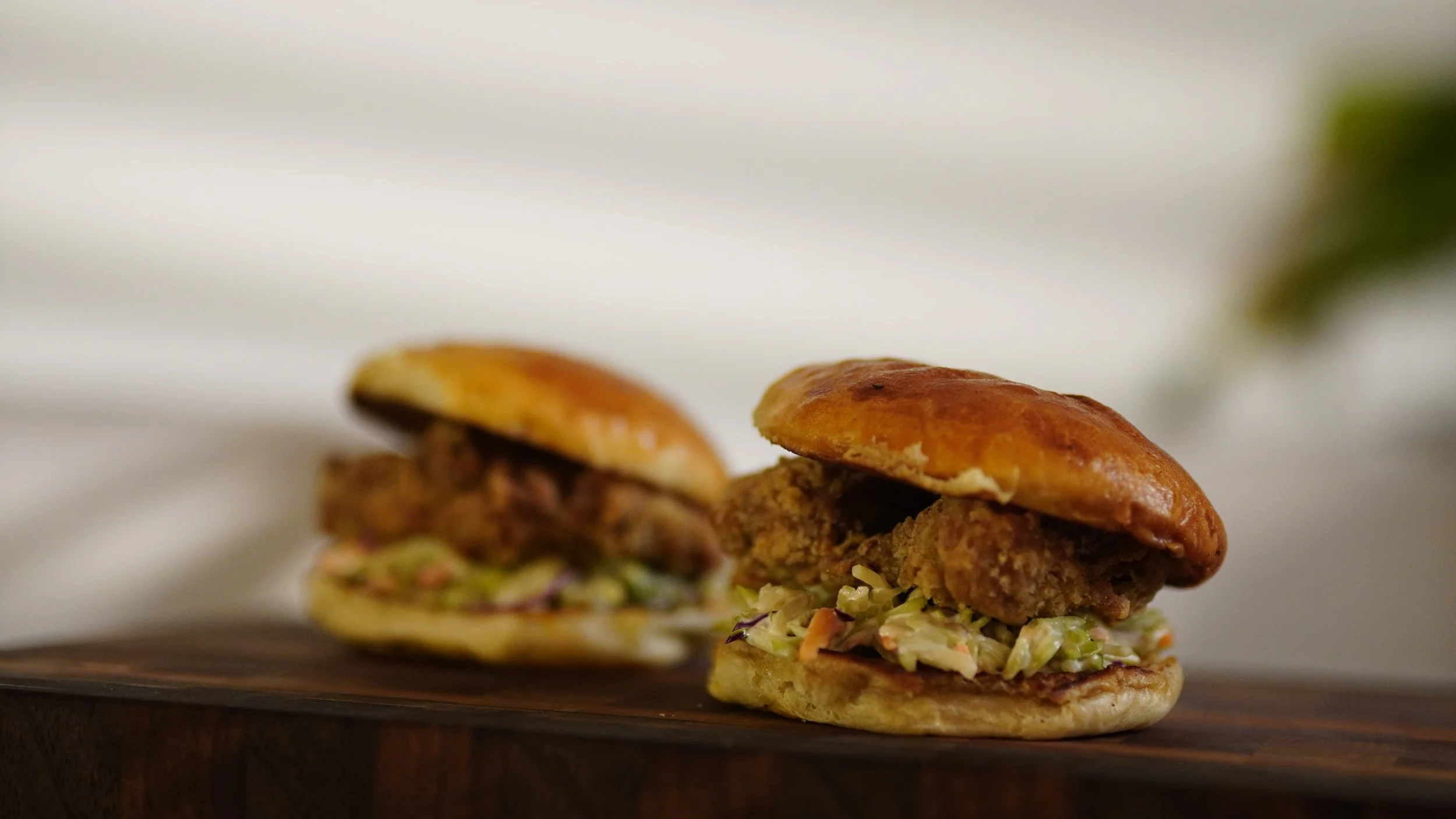 Two fried chicken sandwiches with coleslaw on buns placed on a dark wooden surface.