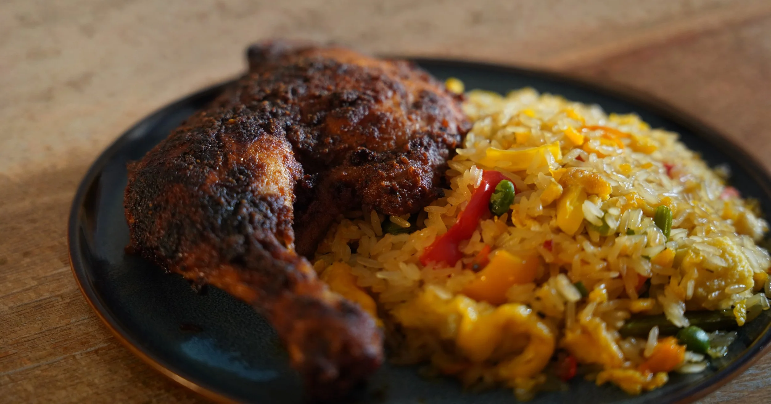 Close-up of a black plate with seasoned roasted chicken and fried rice with vegetables on a wooden surface.