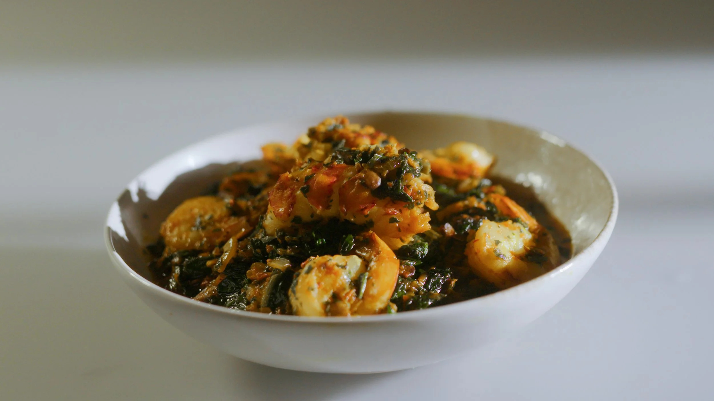 A white bowl filled with a spicy seafood dish, featuring shrimp, vegetables, and herbs.
