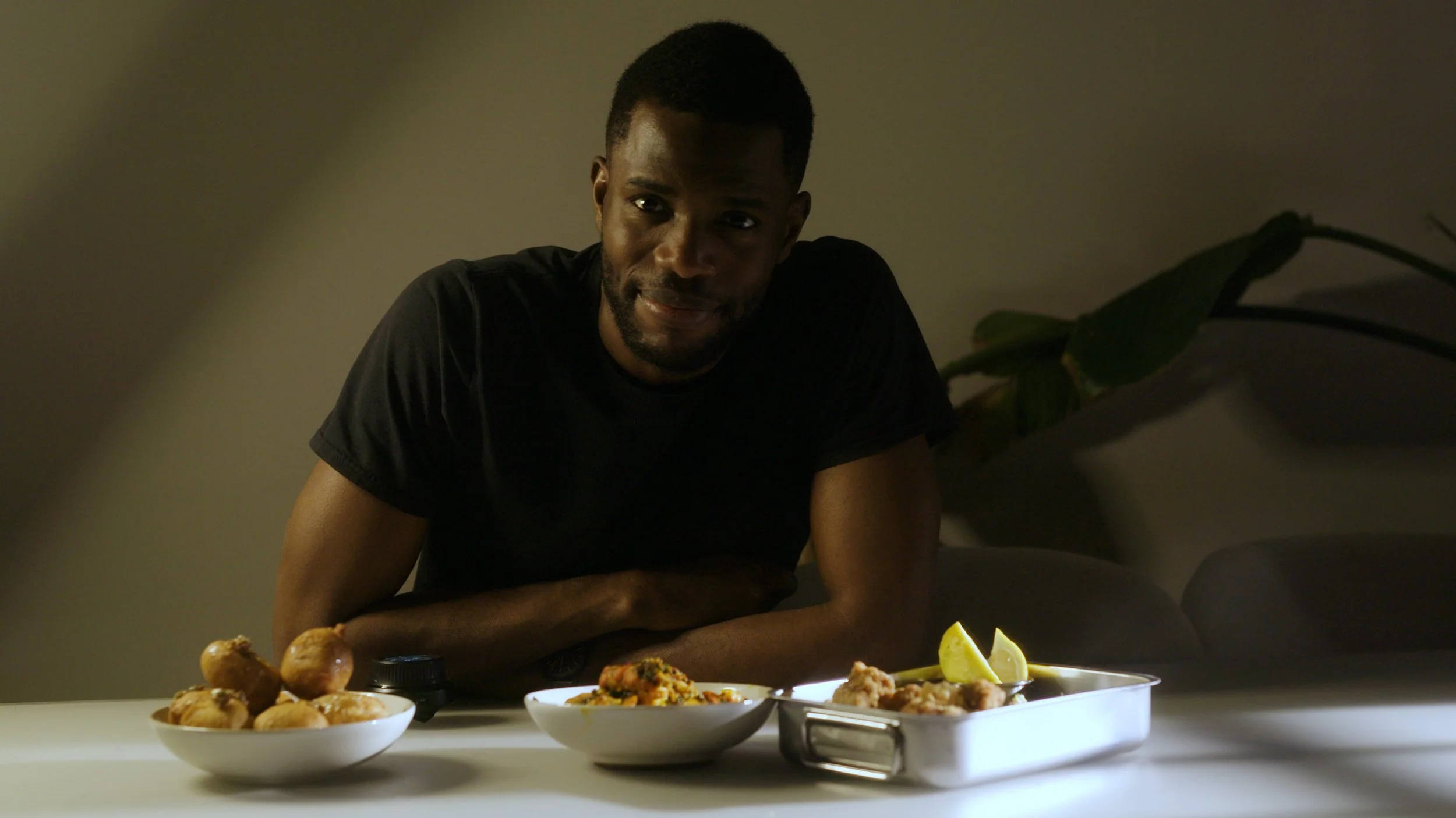 A man with dark skin and short black hair leaning on a table with three plates of east asian and west african food, in a dimly lit room.