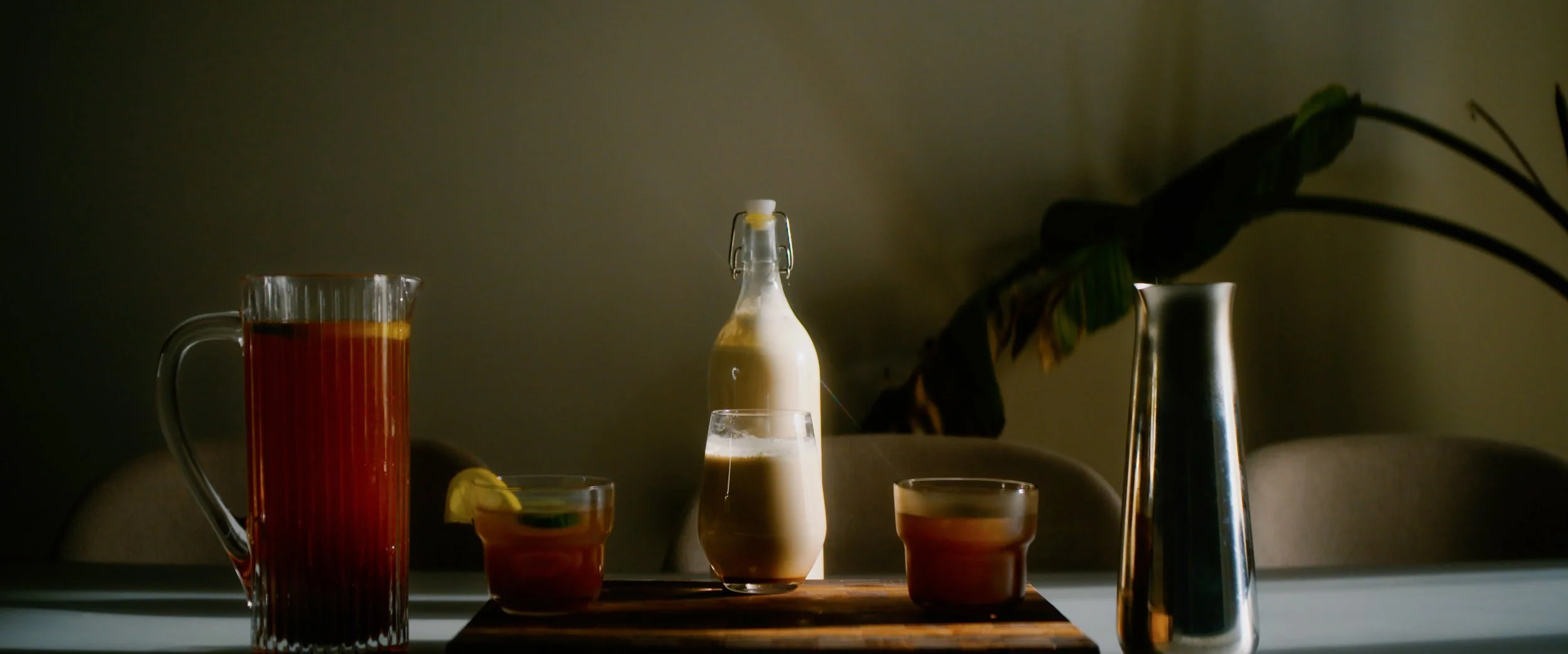 A table with a tall glass pitcher of dark red beverage, two small glasses with lemon wedges, a cream-colored bottle, and a silver vase with large green leaves, lit by natural light