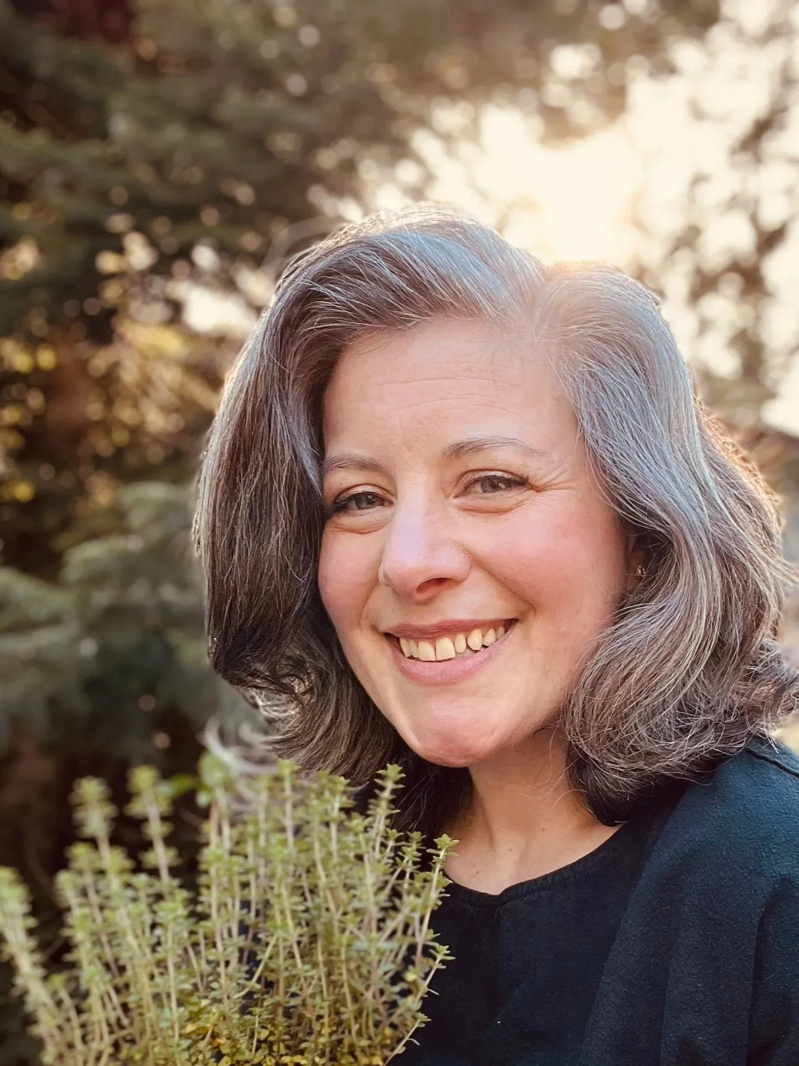 A woman with graying hair smiling outdoors near some thyme, with sunlight filtering through trees in the background.