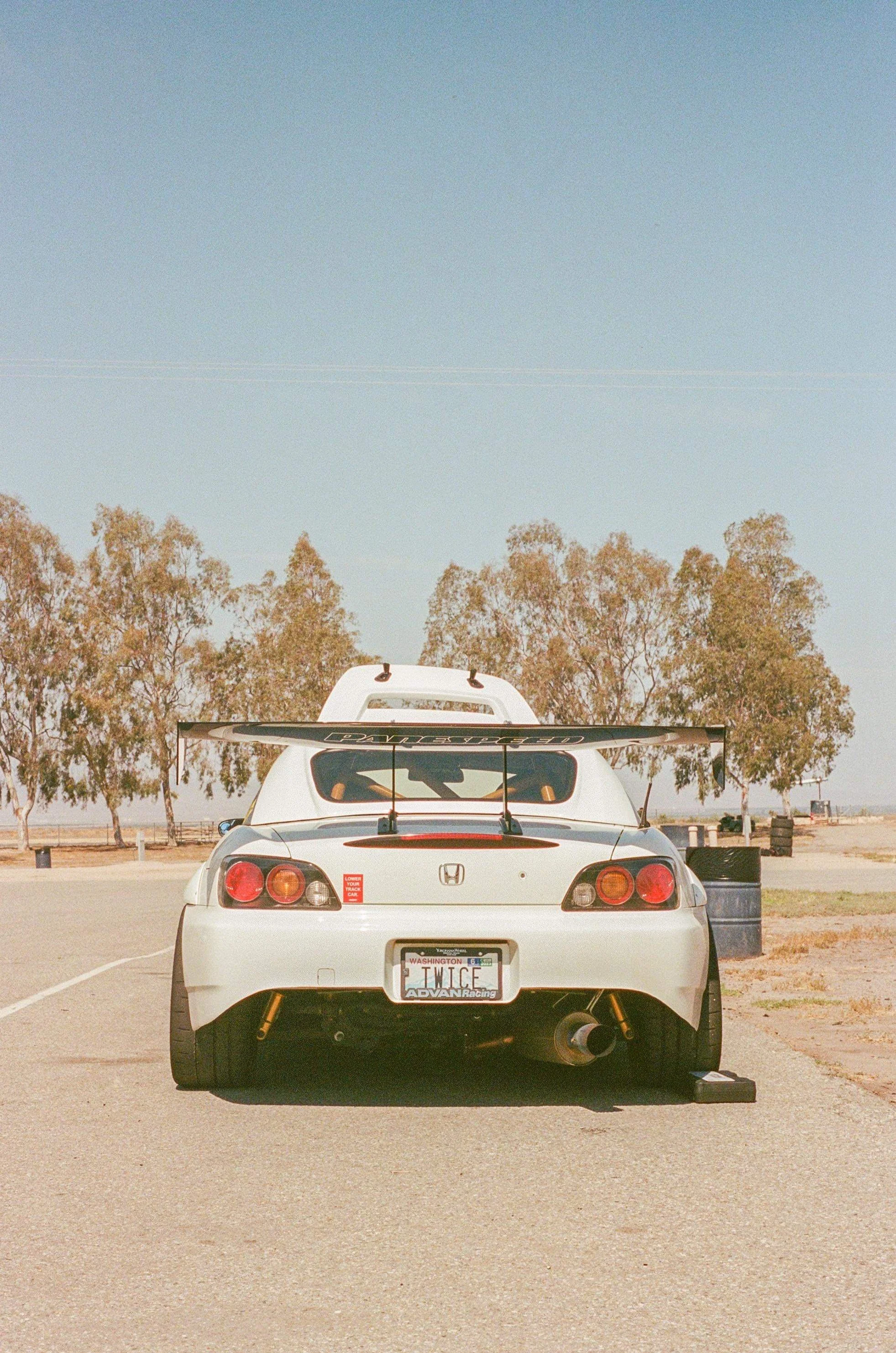 White Honda sports car with a large rear wing, racing decals, and a Washington license plate reading "TWICE" parked on an empty road with trees and a clear blue sky in the background.