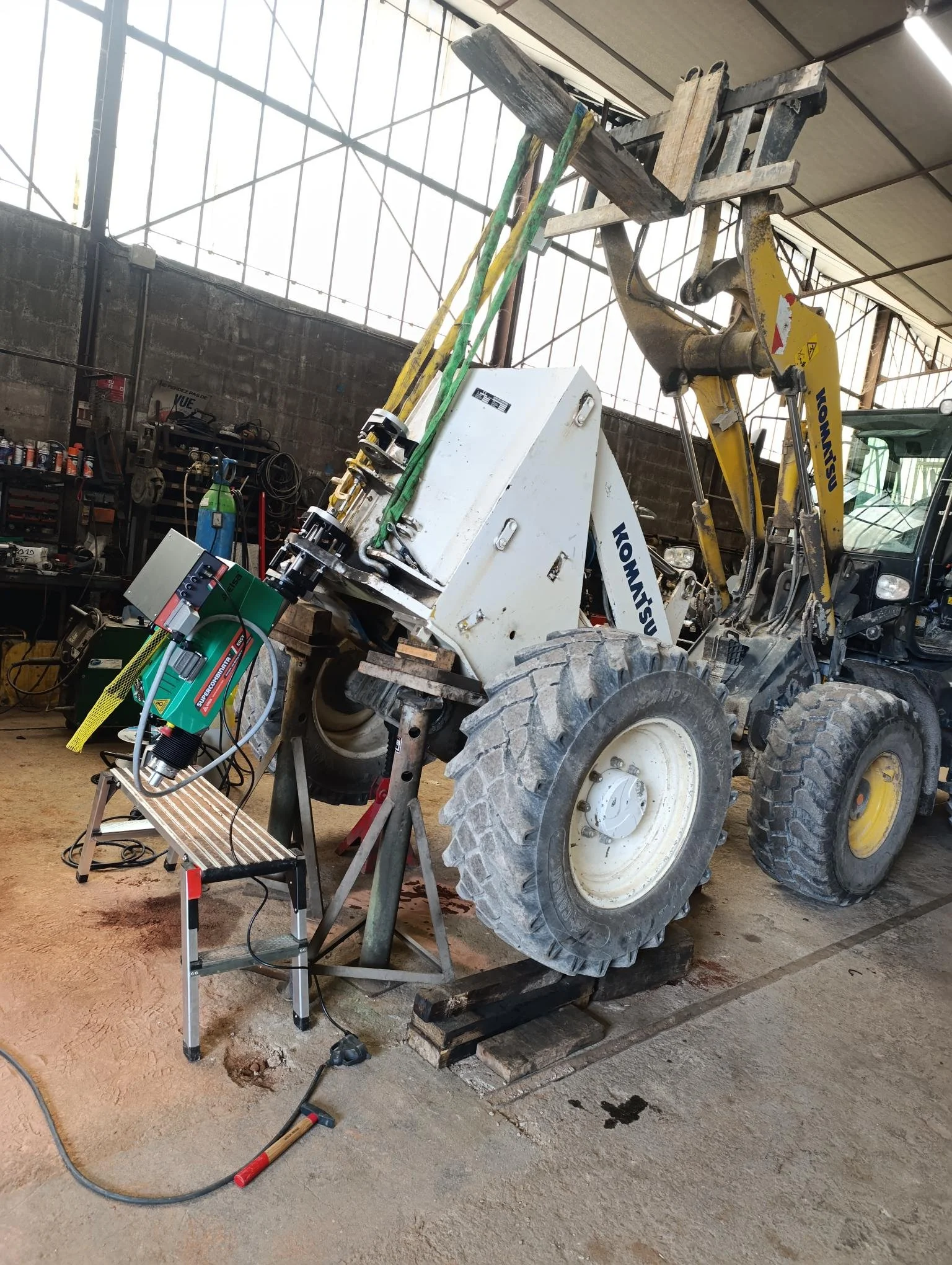 Un chantier ou atelier avec un engin de construction, une machine de soudure, et divers outils de travail sur un sol en terre. Le fond montre un mur en parpaings et des fenêtres en verre.
Alésage portatif, rechargement, réparation chape