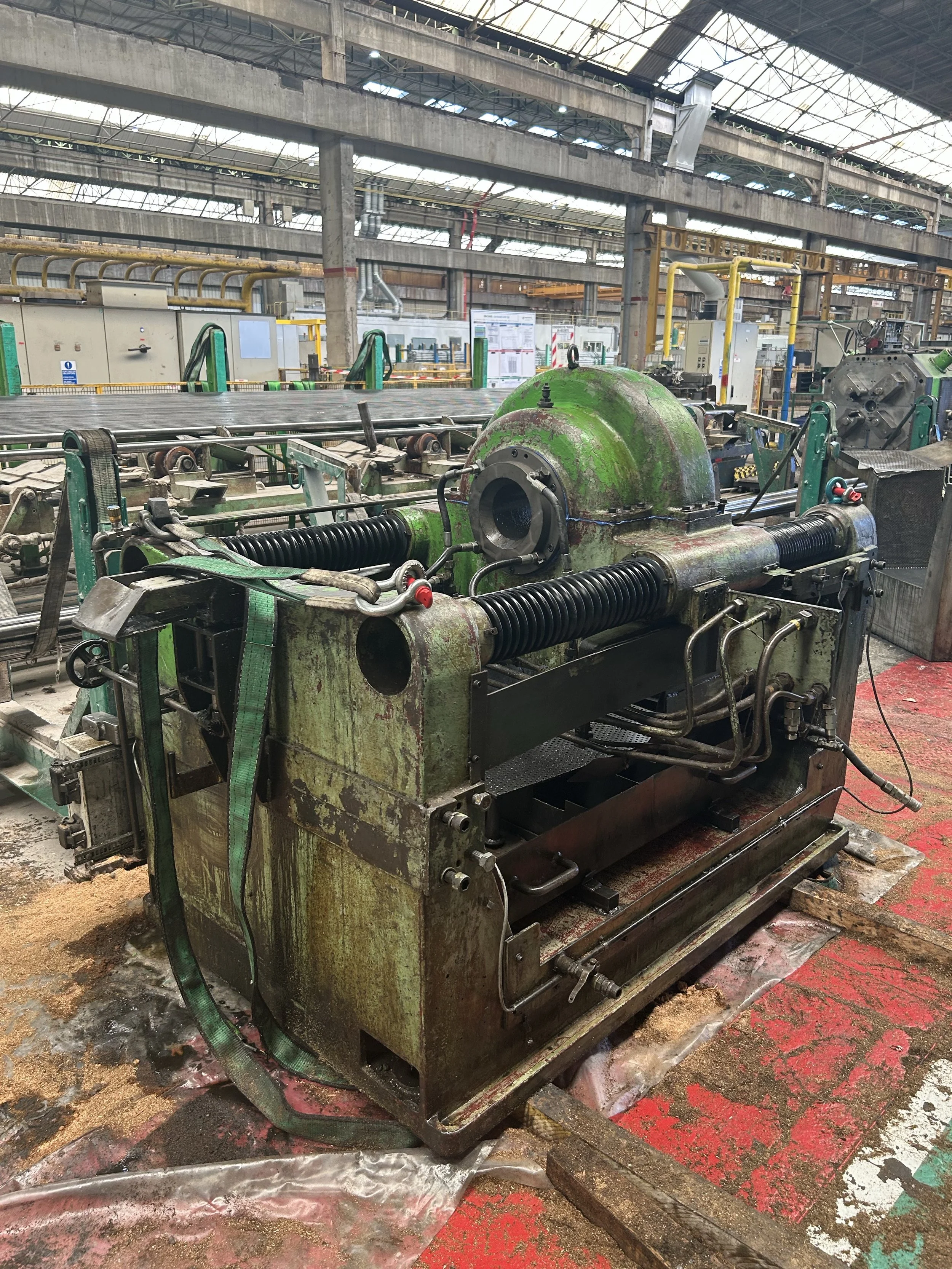 Une machine industrielle ancienne et rouillée dans une usine, avec des tuyaux et des composants mécaniques, sur un sol sale, sous une grande structure en acier.