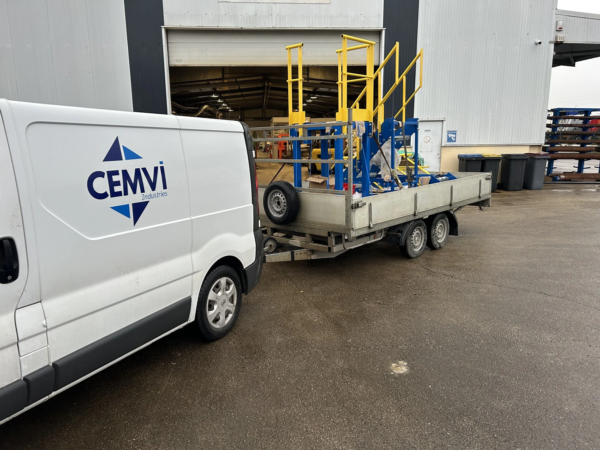 Fichier de transport avec échafaudages jaunes et bleus, stationné devant un bâtiment industriel, avec une voiture blanche à côté portant le logo CEMVI Industries.