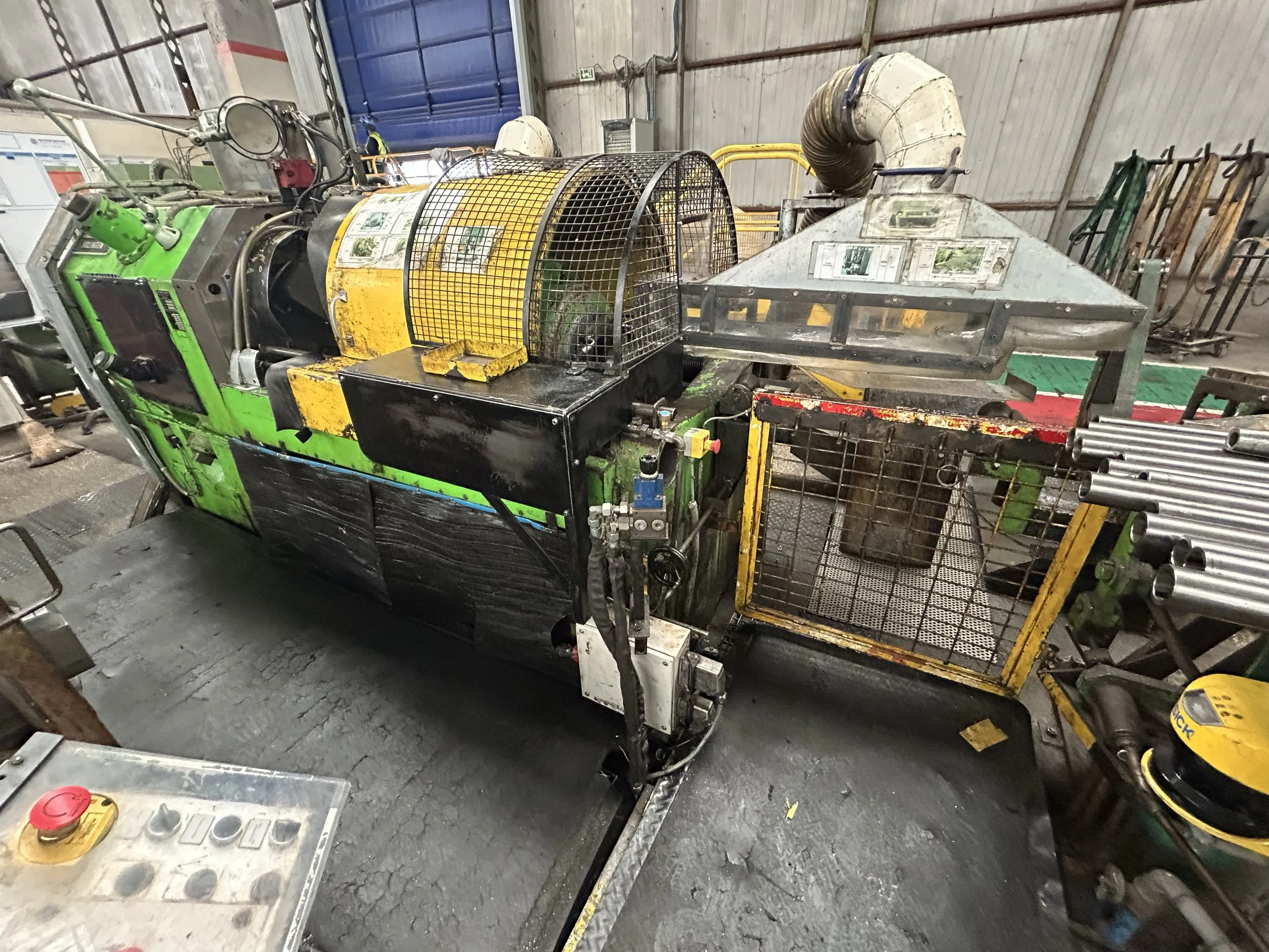 Une machine industrielle ou robotique dans un entrepôt ou un atelier, avec un panneau de contrôle et plusieurs pièces métalliques ou tubes à proximité.