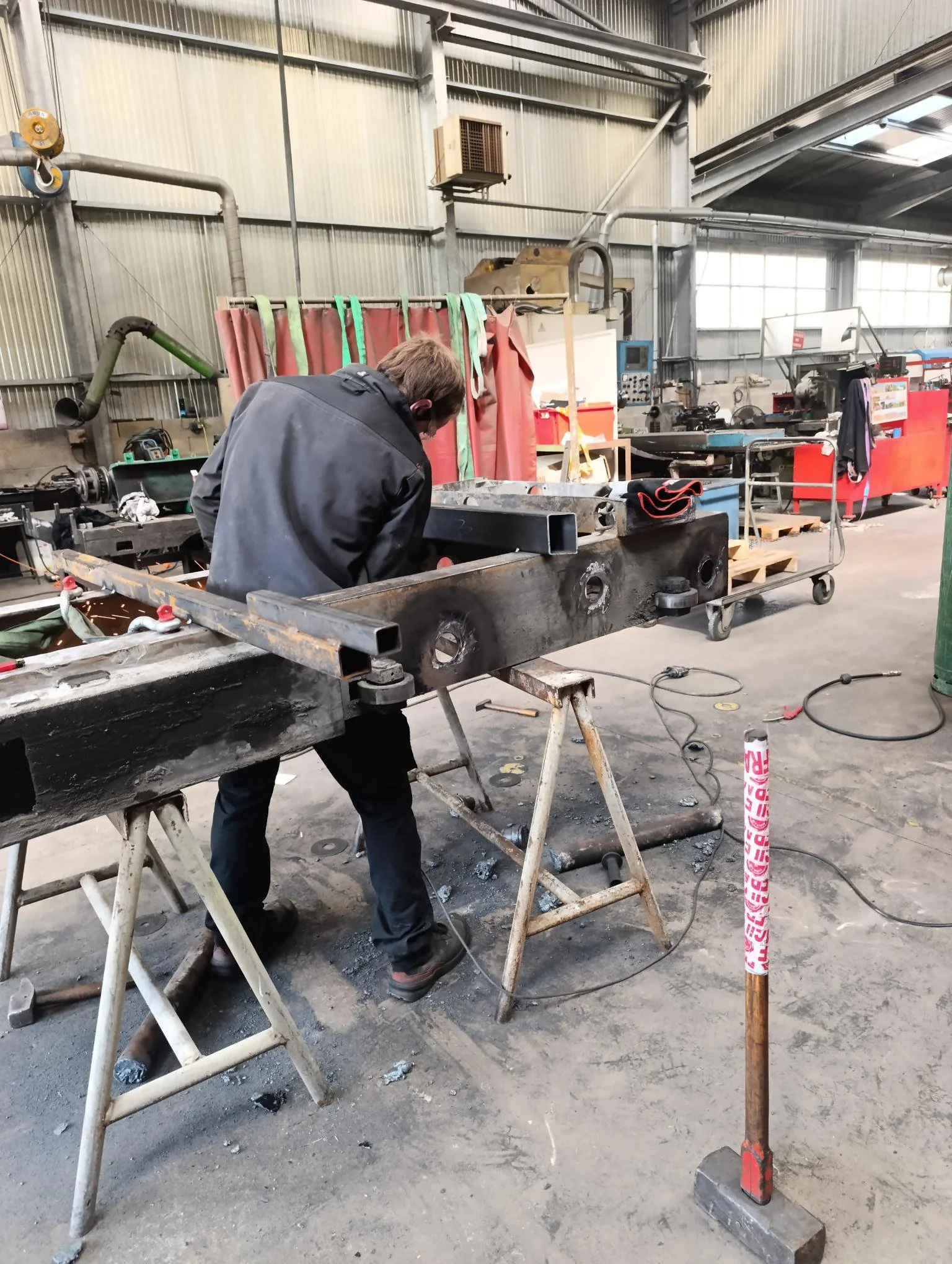 Un homme travaillant dans un atelier de fabrication ou de réparation, utilisant des outils sur une pièce métallique. L'atelier est spacieux avec des murs en métal, des équipements industriels, et des bacs de rangement.