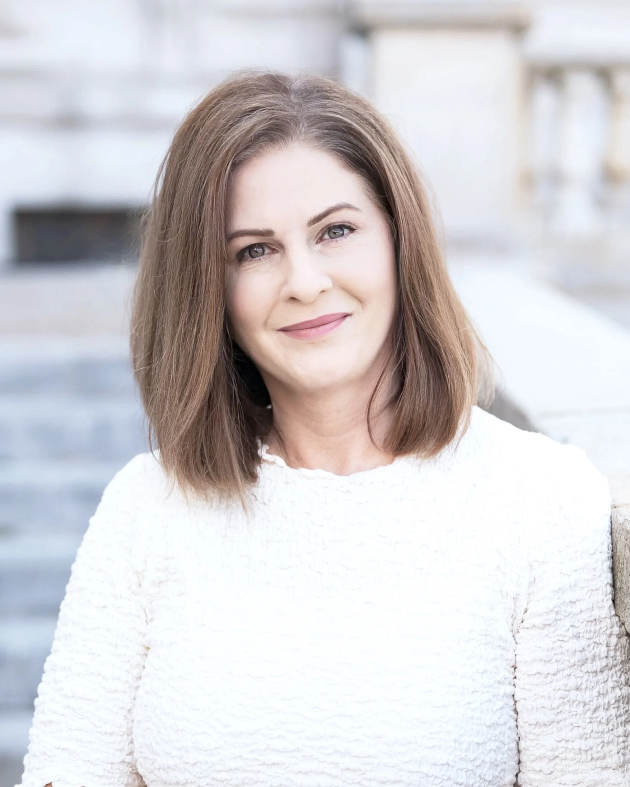Portrait of a woman with shoulder-length brown hair smiling, wearing a white sweater, outdoors near a white brick wall.