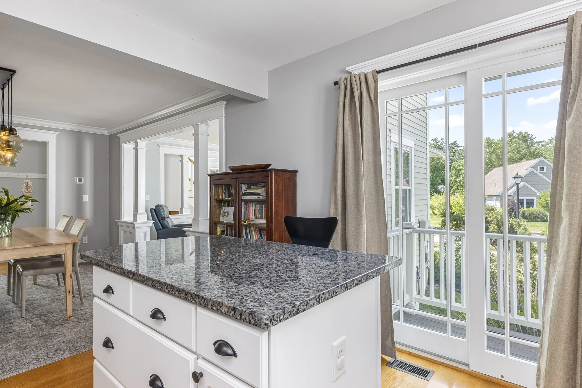 Bright kitchen area with large granite-topped island, wooden dining table, and large windows with beige curtains overlooking a backyard with trees and houses.