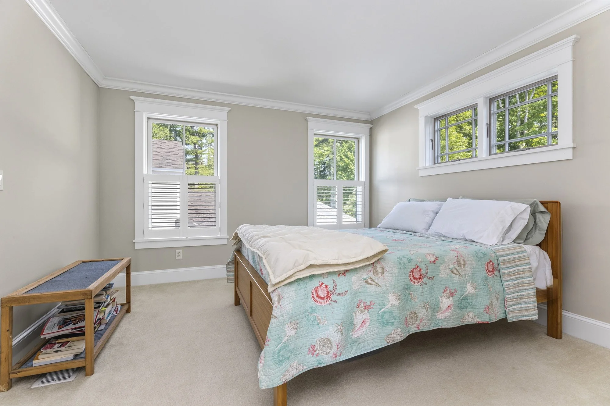 A bedroom with a bed covered with a floral quilt, three white pillows, and a duvet, next to windows with white frames and greenery outside. There is a small wooden nightstand with a book and magazines, and a beige carpet on the floor.