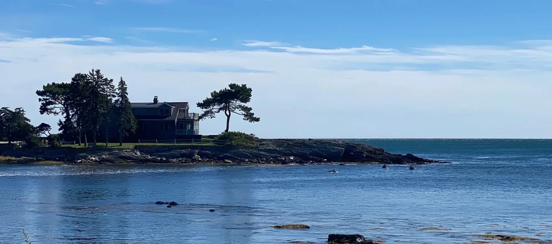 A house on a rocky shoreline with trees nearby, overlooking the ocean, under a partly cloudy sky.