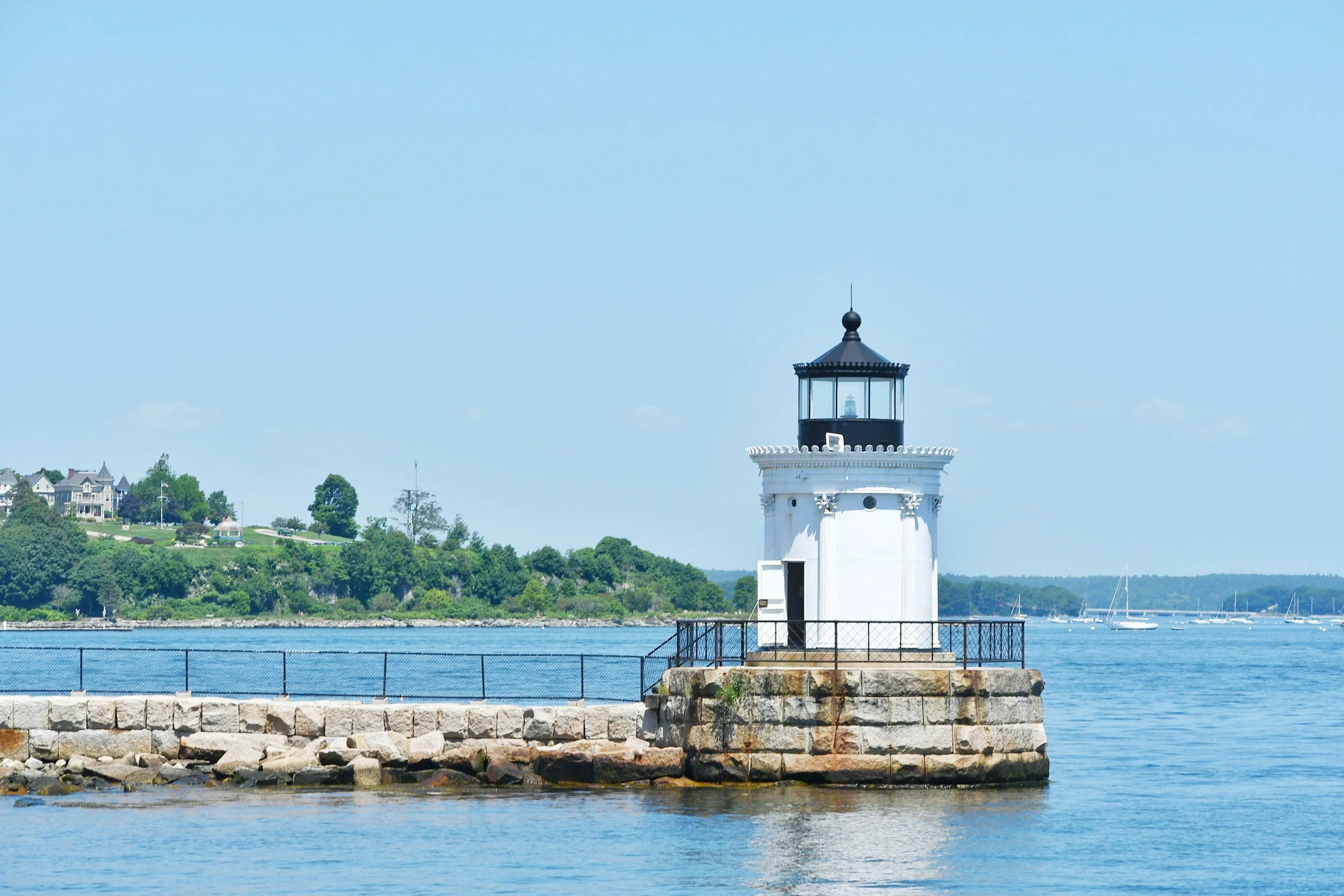 A white lighthouse with a black top and glass windows, situated on a stone-paved pier by the water. In the background, there's a hillside with houses and green trees, and sailboats are visible on the water under a clear blue sky.