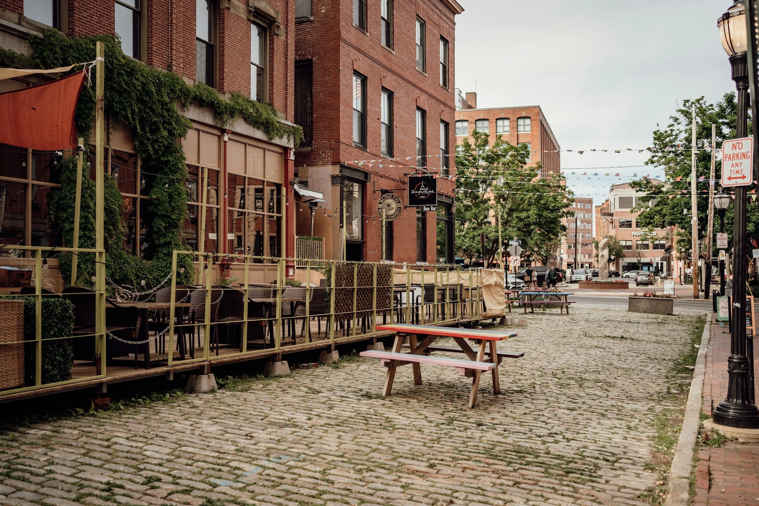 Empty outdoor dining area with tables and chairs in front of a red brick building on a cobblestone street, with trees and hanging string lights.