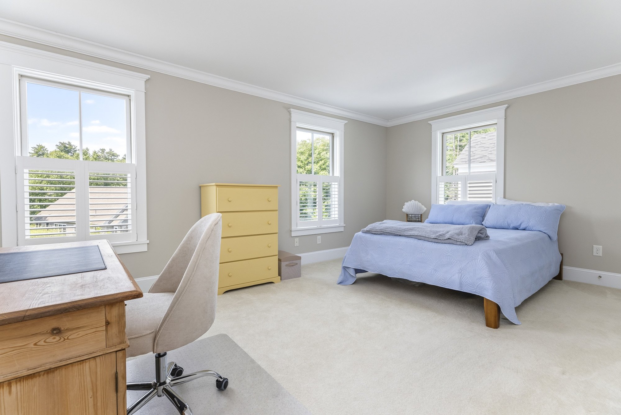 A bedroom with three windows, a bed with a blue bedspread, a yellow dresser, a desk with a chair, and natural light coming through the windows.